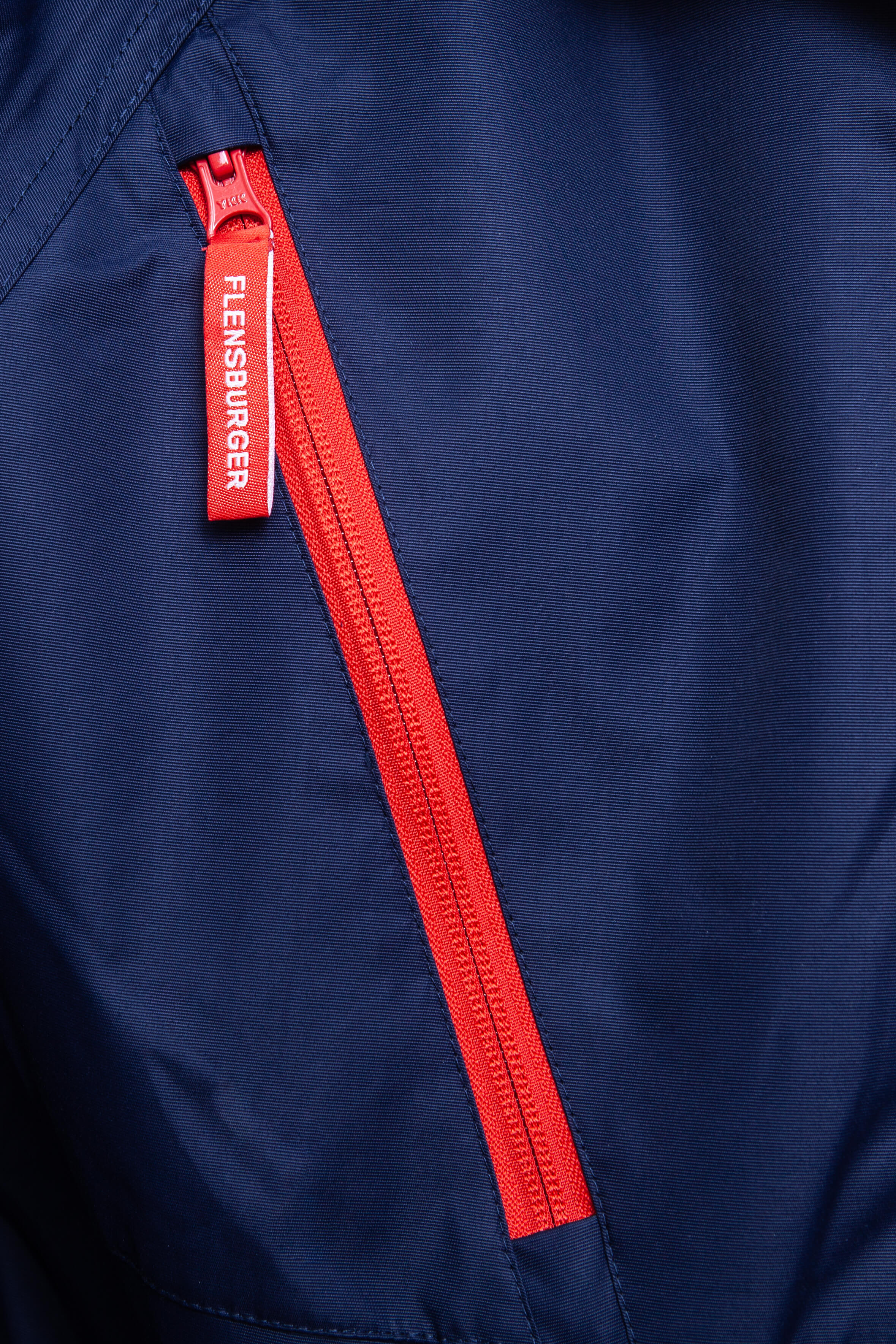 Detailaufnahem eines roten Reißverschluss auf navyblauem Parka mit rotem Zipperanhänger, darauf steht in weißen Buchstaben "FLENSBURGER".