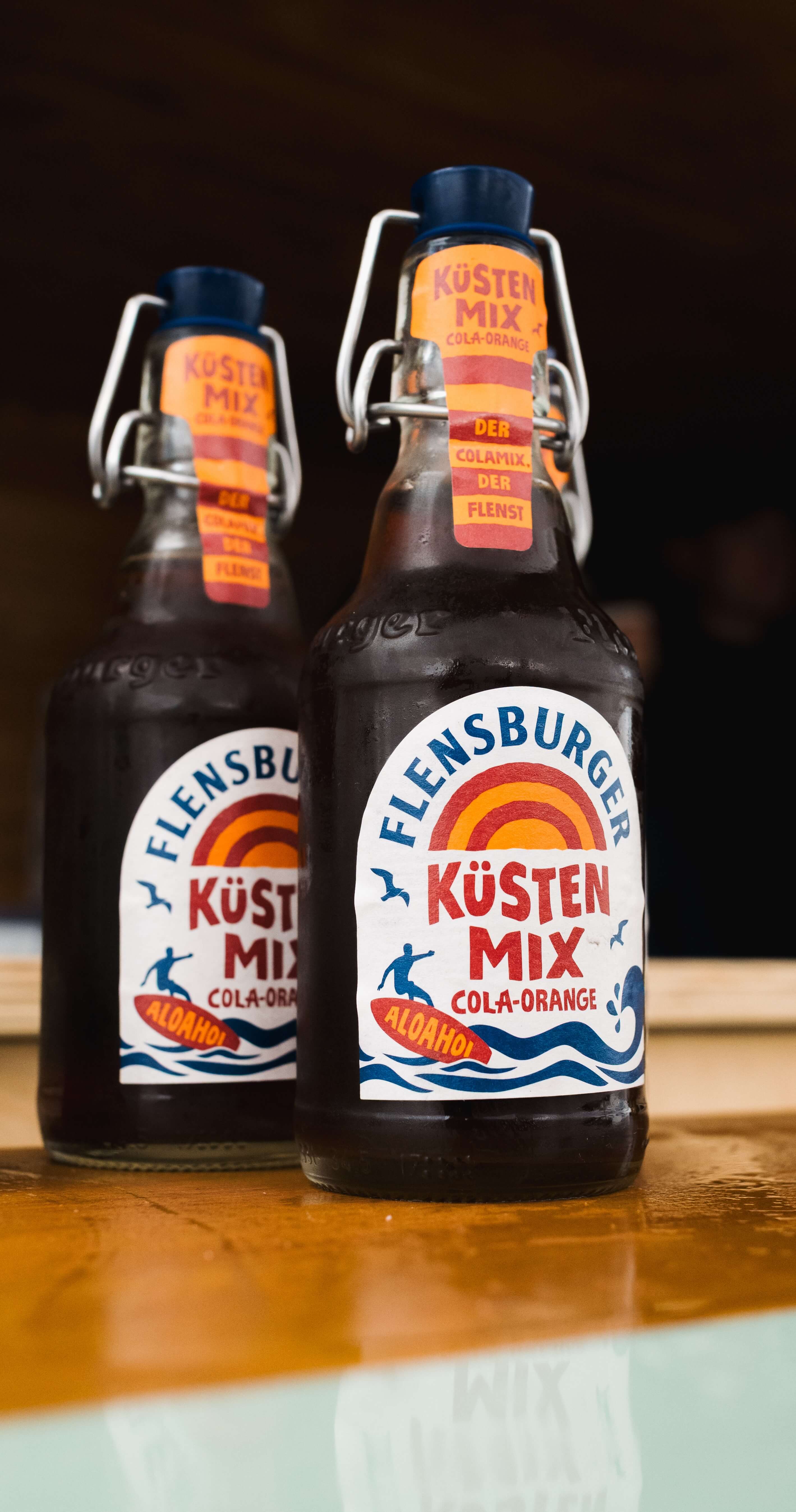 Flensburger Küstenmix "Cola-Orange" in braunen Mehrwegflaschen steht auf einem Holztisch. Die Flaschen haben Etiketten mit buntem Design und beschreiben das Produkt sowie den Alkoholgehalt von 0,0% vol.
