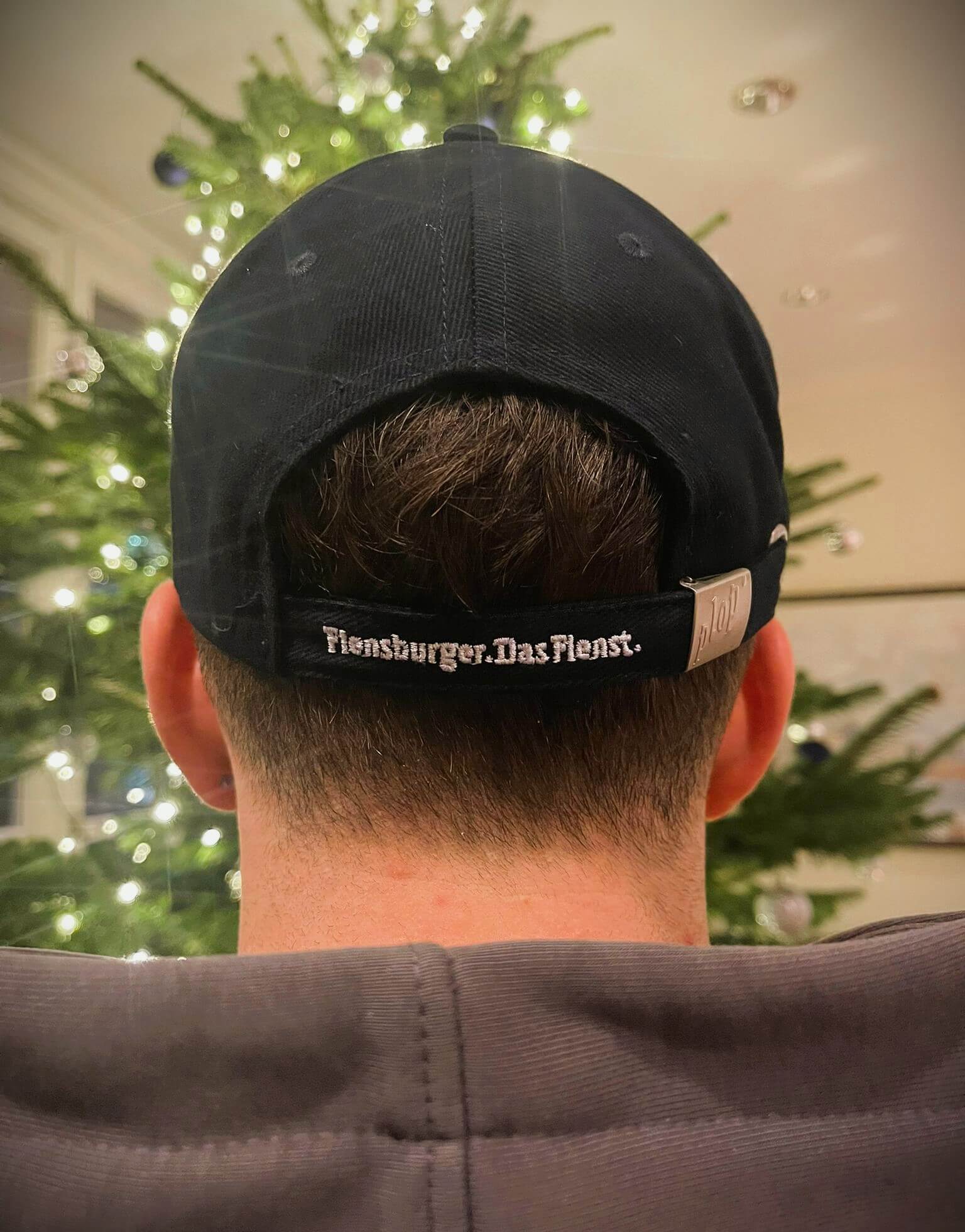 Mann von hinten mit kurzen Haaren trägt eine dunkelblaue Baseballcap mit der weißen Stickerei „Flensburger. Das Flenst.“ auf dem verstellbaren Riemen. Im Hintergrund leuchtet ein geschmückter Weihnachtsbaum.