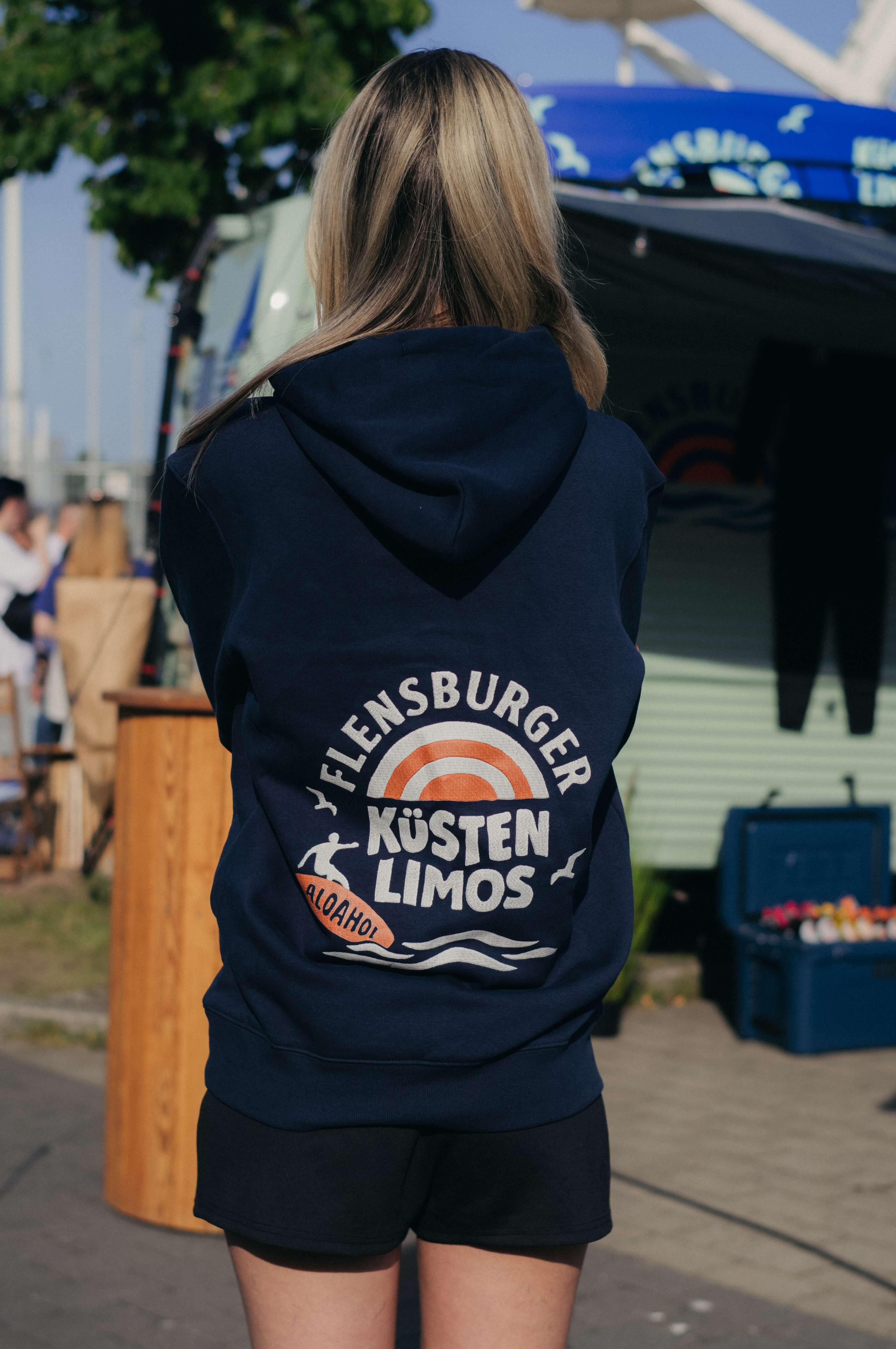 Junge Frau mit Pferdeschwanz trägt einen navyblauen Hoodie mit dem Logo "Flensburger Küstenlimos" auf dem Rücken. Im Hintergrund stehen Stände und Bäume sind sichtbar.