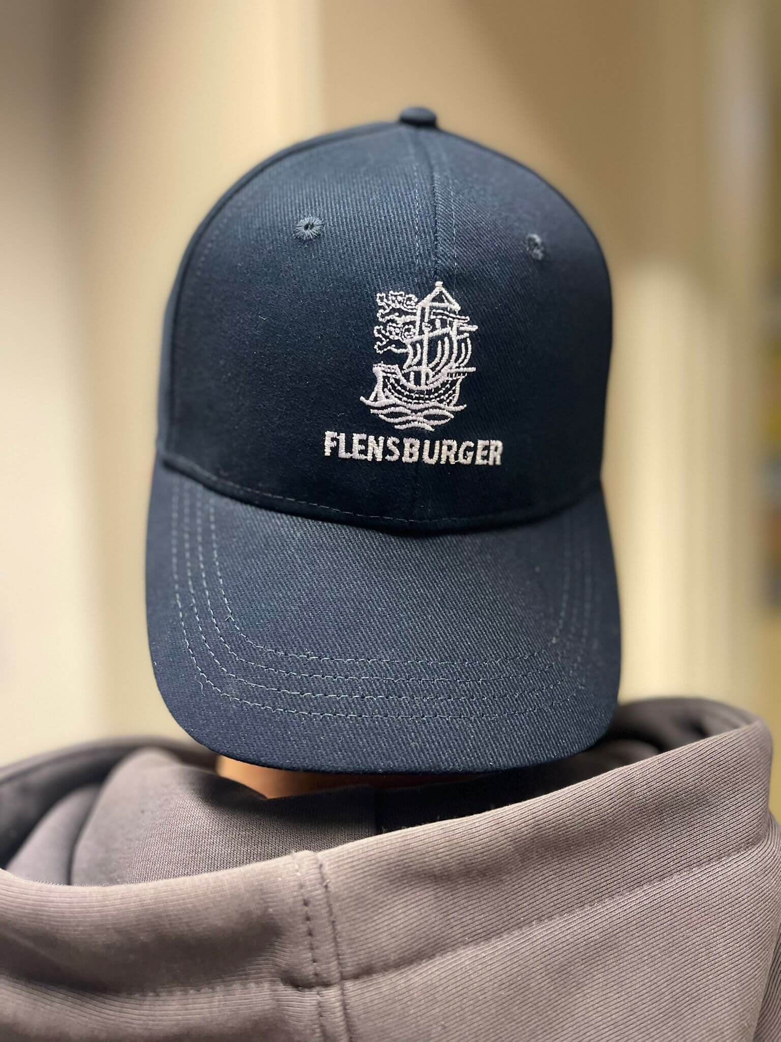 Eine dunkelblaue Cap mit einem weißem Schiff-Logo und dem Schriftzug „FLENSBURGER“ auf der Vorderseite. Die Cap wird mit dem Schild nach hinten von einer Person mit grauem Kapuzenpulli getragen.