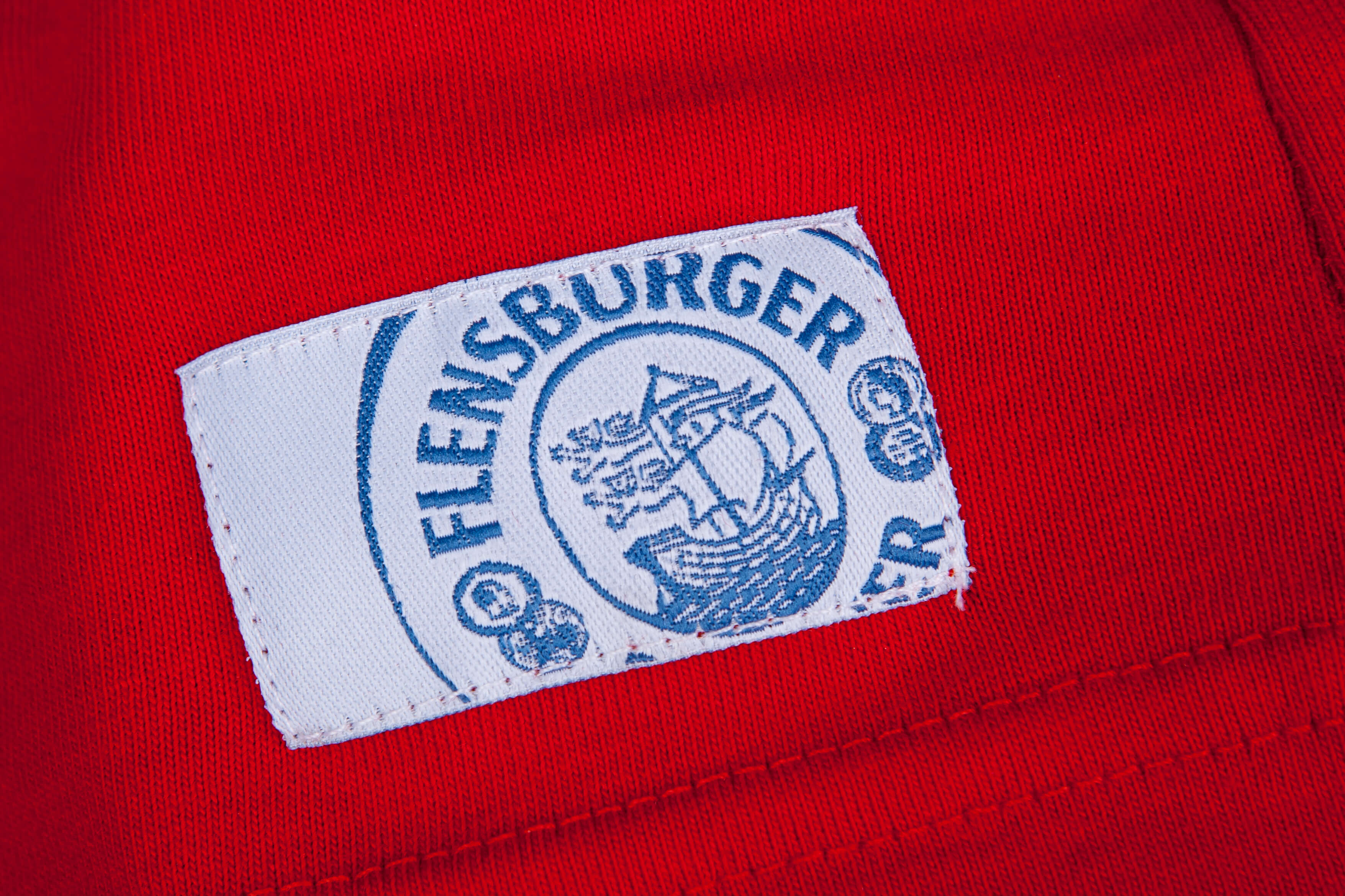 Auf einem roten Baumwollstoff ist ein weißes, rechteckiges Etikett mit dem blauen Schriftzug "FLENSBURGER" und einem Segelschiff in der Mitte angenäht.