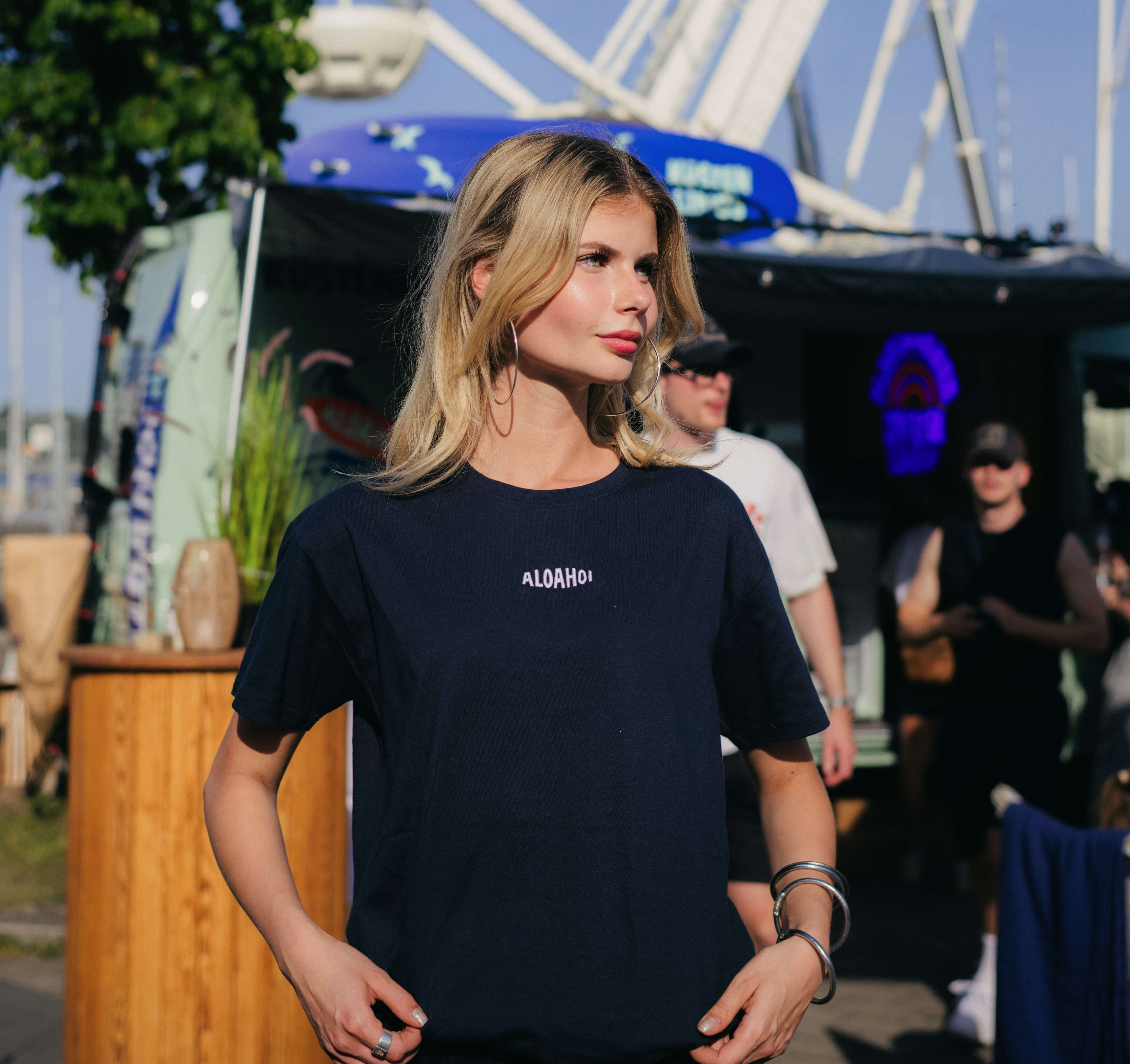 Junge Frau trägt ein dunkelblaues T-Shirt mit der Aufschrift "ALOHAOI". Sie steht im Freien, umgeben von anderen Personen und einem Food-Truck im Hintergrund.