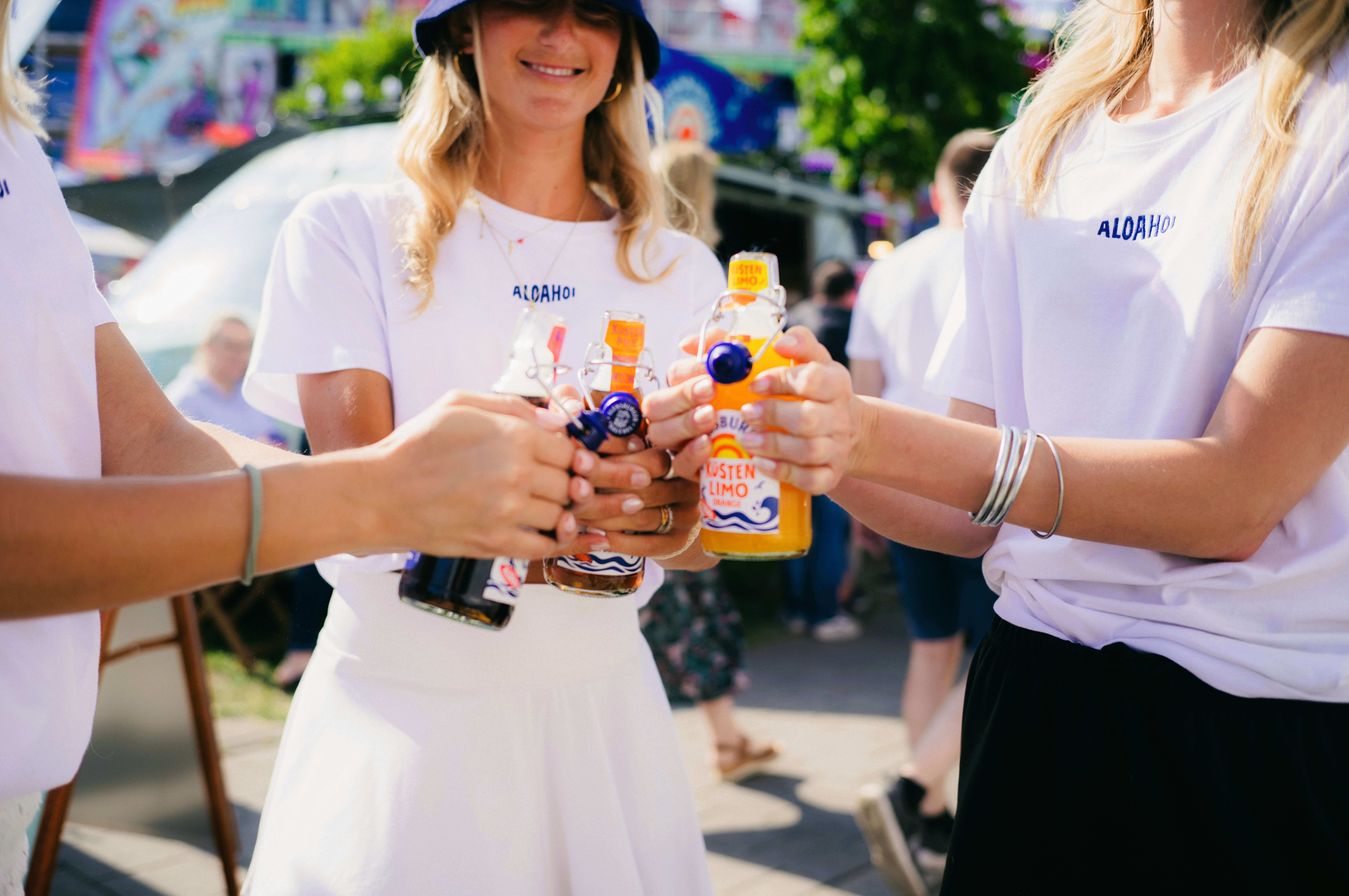 Drei Personen halten Flensburger Küstenlimos in ihren Händen. Sie tragen weiße T-Shirts mit dem Logo „ALOHA“. Hinter ihnen sind Menschen und eine bunte Kulisse zu sehen.