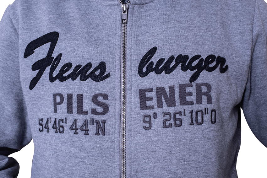 Detailansicht der Kapuzenjacke mit Reißverschluss zeigt die schwarze Aufschrift  „Flensburger“ und darunter in grauer Schrift „PILS 54°46'44''N“ sowie „ENER 9°26'10''O“ als geografische Koordinaten. Die Aufschrift ist auf Brusthöhe angebracht.