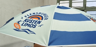Der Sonnenschirm ist in hellblau und weiß gestreift. Er trägt das Logo „Flensburger Küsten Limos“ und ist auf einem Tisch platziert.