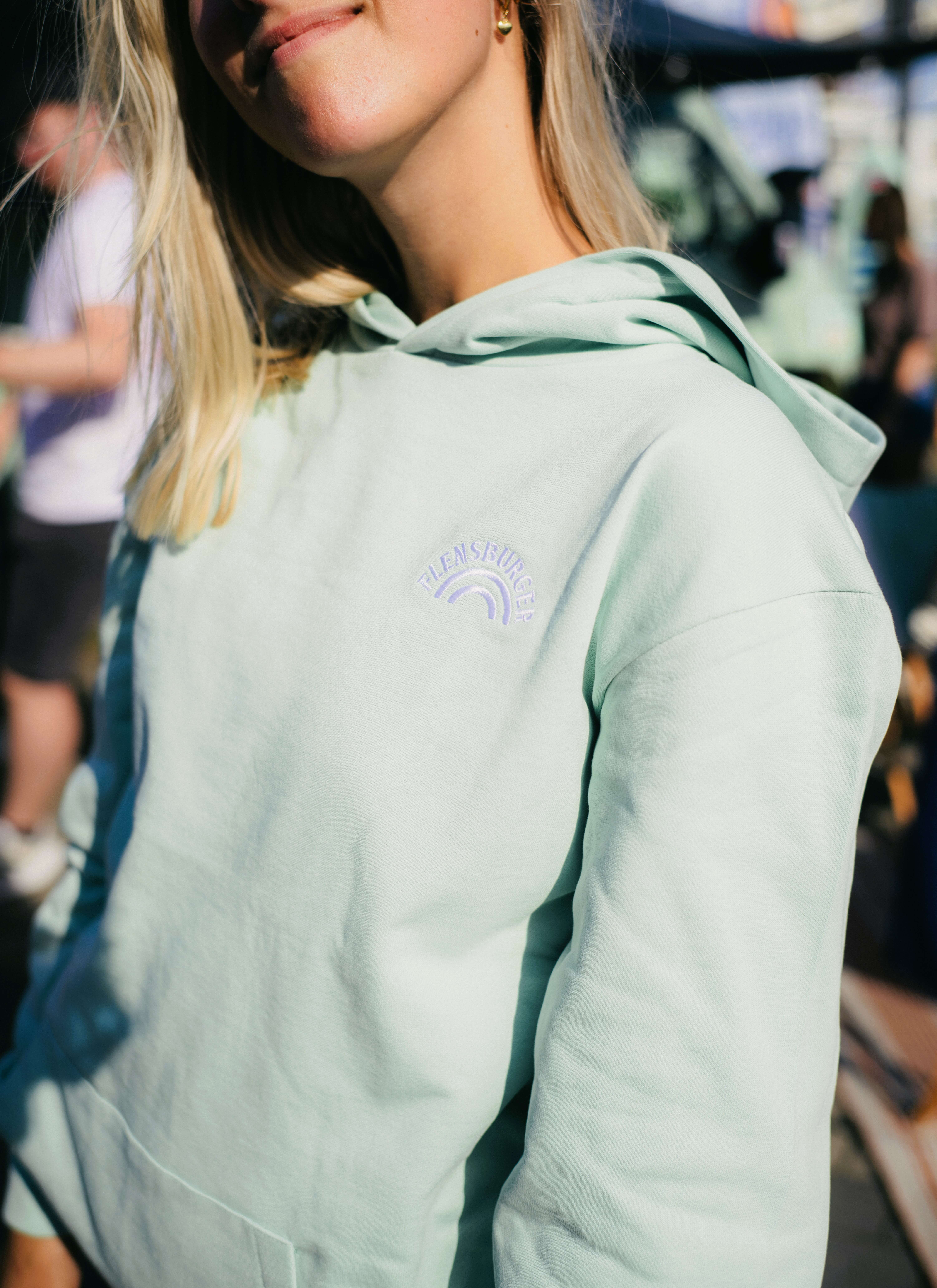 Junge Frau mit blonden Haaren trägt einen mintfarbenen Hoodie mit dem aufgestickten Logo "FLENSBURGER". Sie lächelt leicht und steht in einer belebten Umgebung.