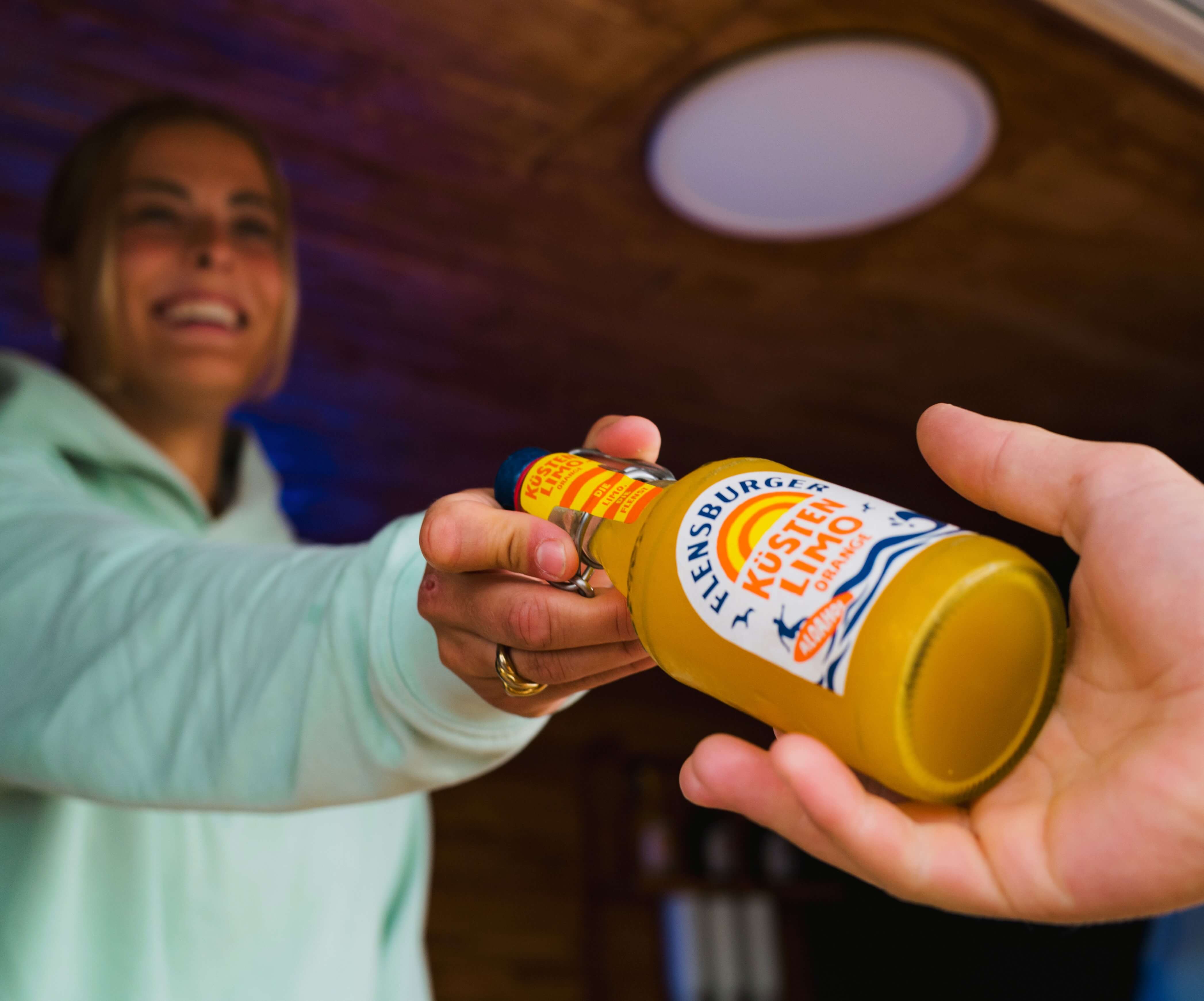 Eine Frau lächelt, während sie eine Flasche Flensburger Küstenlimo „Orange“ mit Alk. 0,0% in der Hand hält und sie an eine andere Person übergibt.