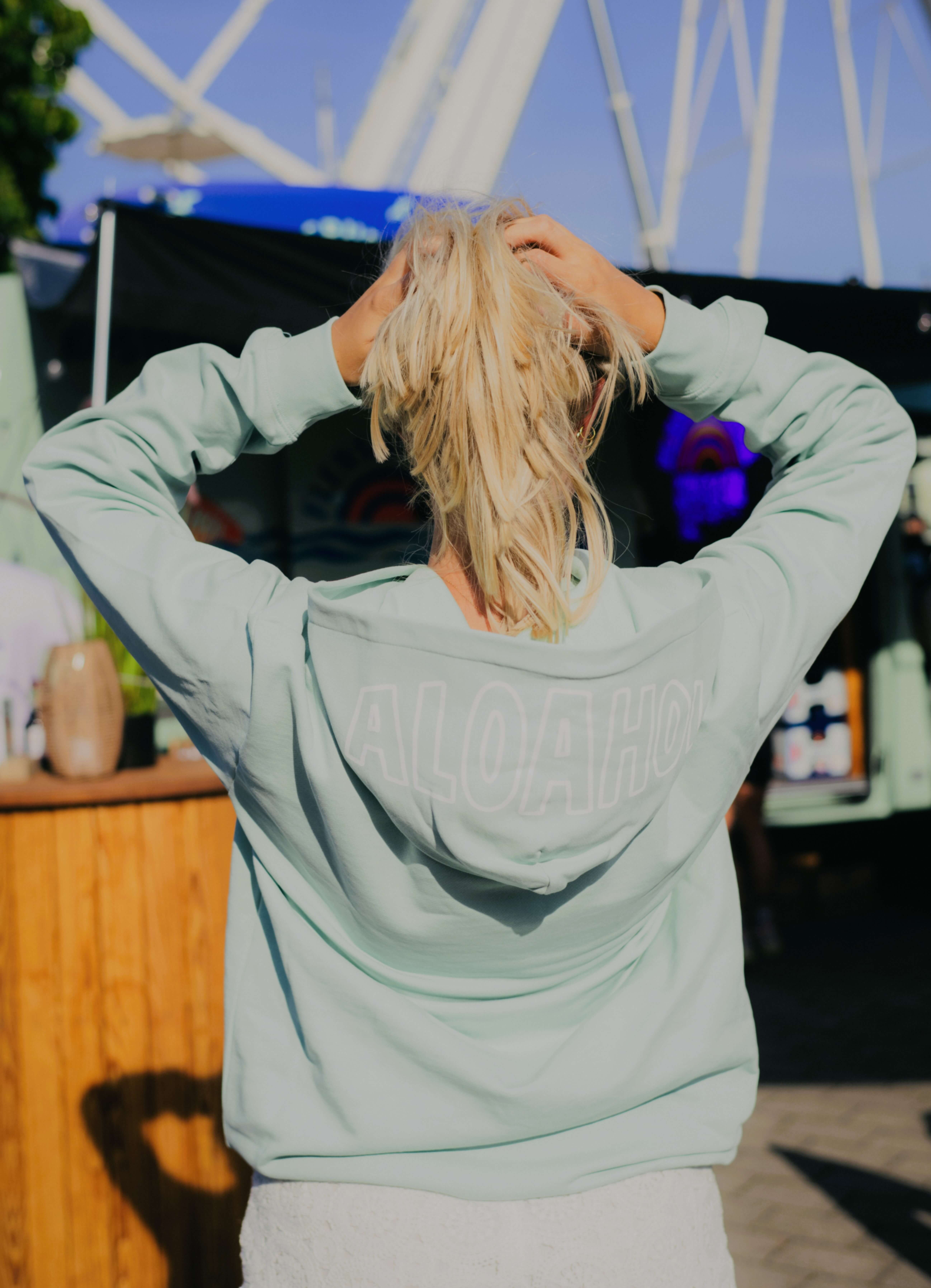 Frau mit blonden Haaren steht mit dem Rücken zur Kamera und hält ihr Haar mit beiden Händen hoch. Sie trägt einen mintgrünen Hoodie mit dem Aufdruck "ALOAHOI".