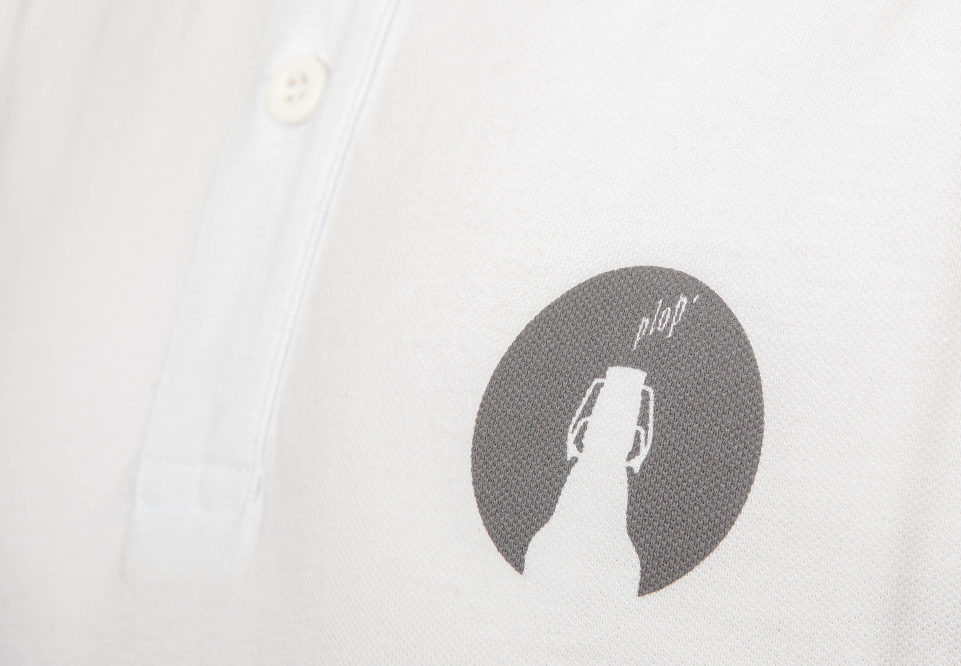 Detailaufnahme vom Aufdruck eines weißen Polo-T-Shirt auf der brust. Es zeigt einen dunkelgrauen Kreis in dem eine Bügelverschlussflasche und der Schriftzug „plop“ zu sehen`ist.
