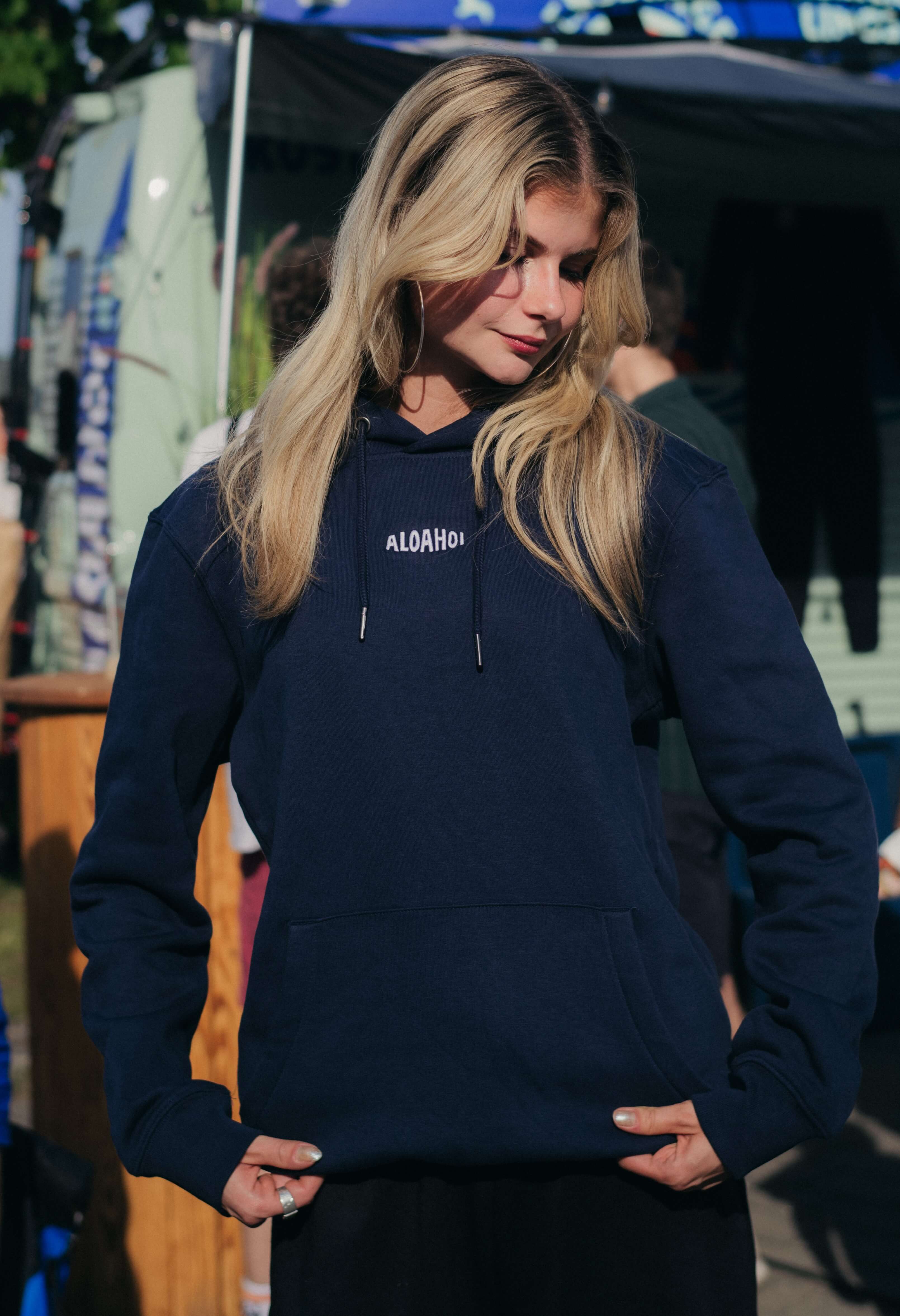 Junge Frau mit langen blonden Haaren trägt einen dunkelblauen Kapuzenpullover mit dem Schriftzug „ALOHAH“ und posiert während eines sonnigen Außenevents.