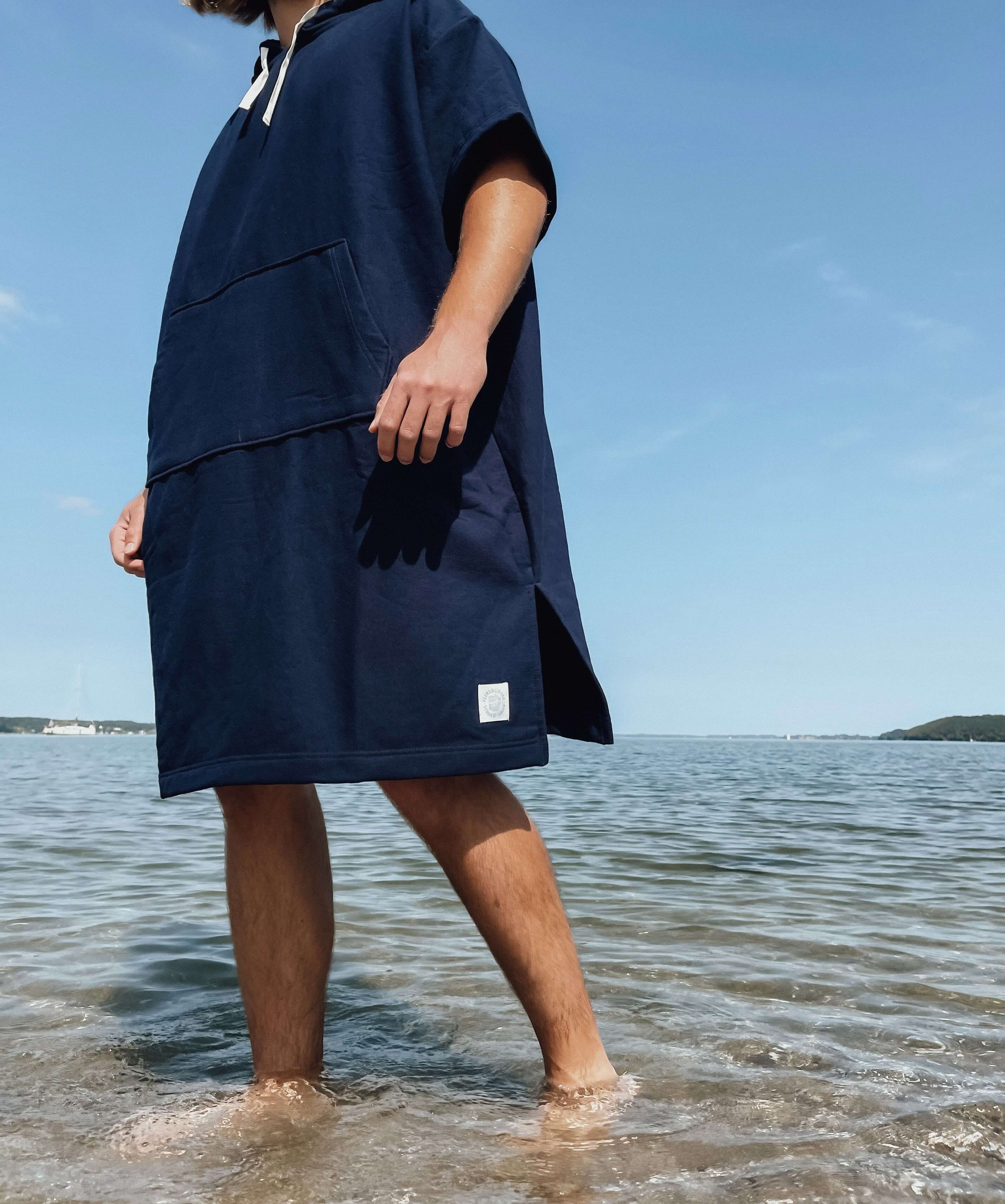 Person steht mit nackten Beinen im flachen Wasser am Strand und trägt einen blauen kurzärmligen Strand-Poncho mit großer Tasche vorne und weißem Logo unten am Saum.