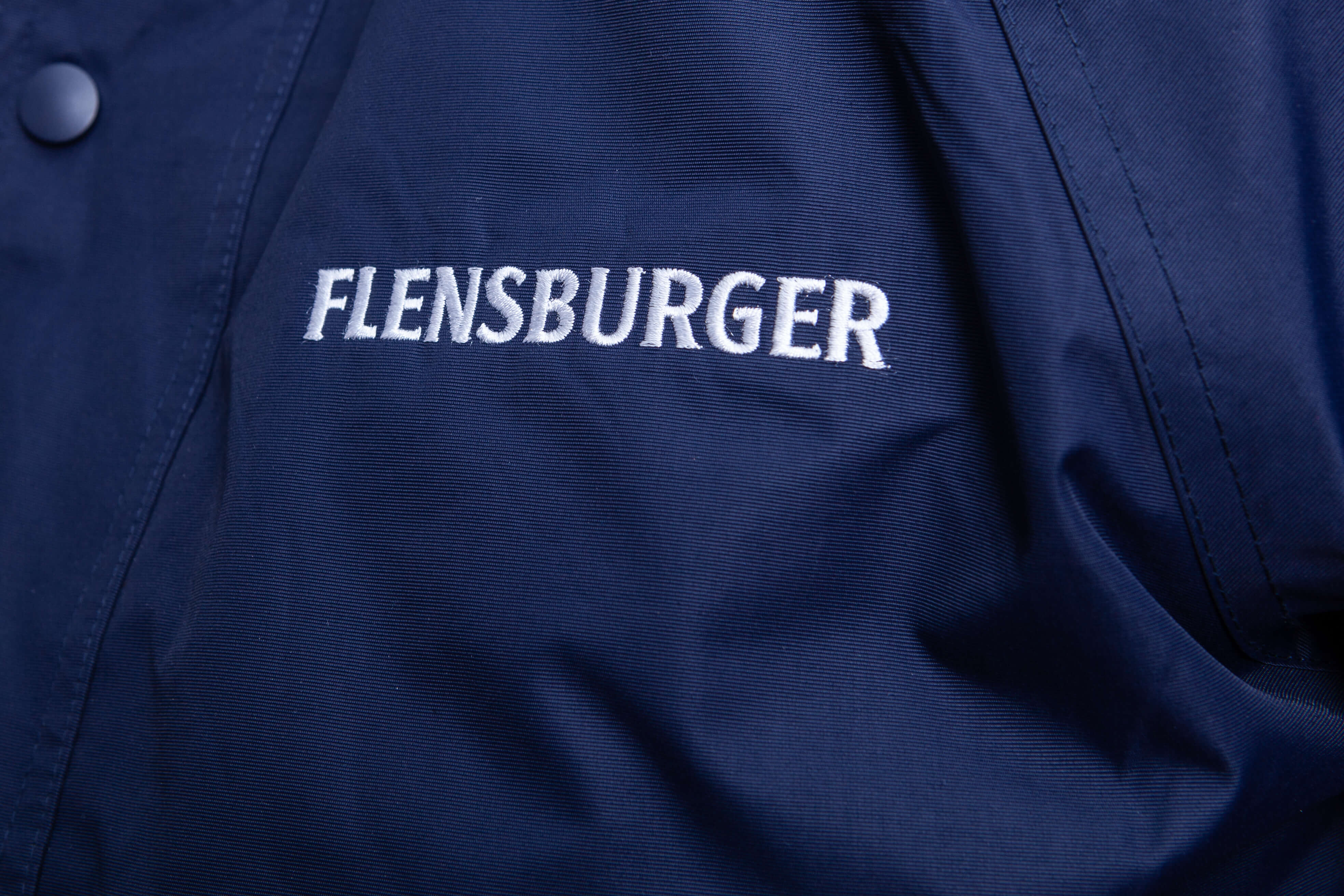 Detailaufnahme eines dunkelblauen Parkas mit der weißen Stickerei "FLENSBURGER" auf der Brust.