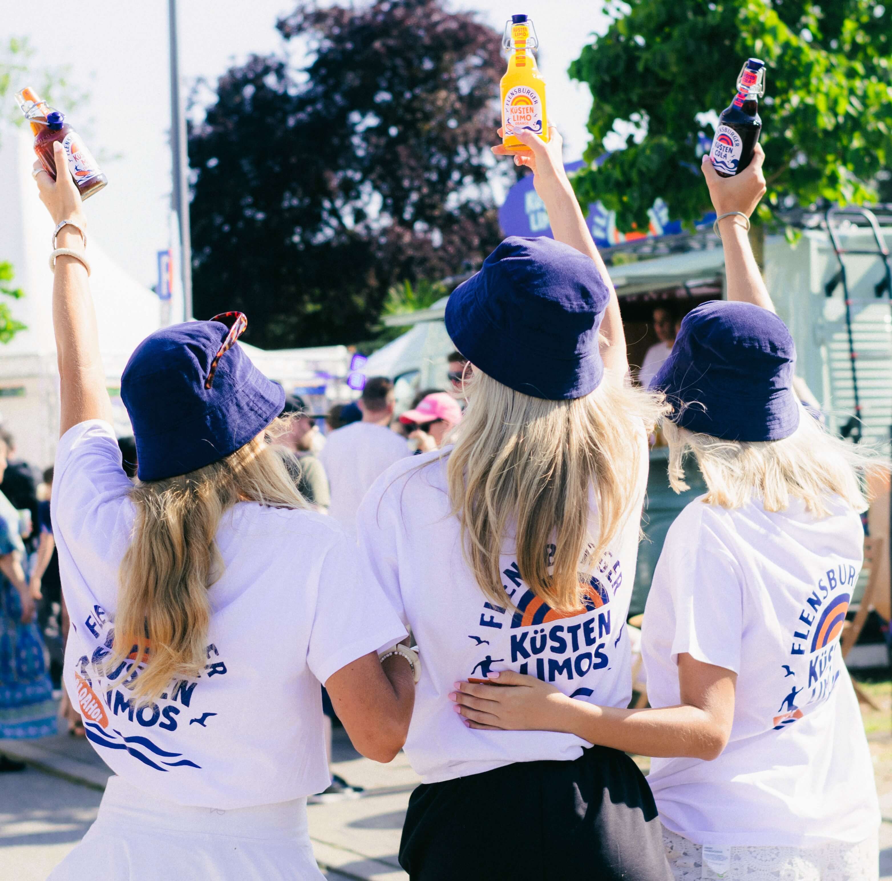 Drei Frauen tragen weiße T-Shirts mit dem Logo "Flensburger Küstenlimos" auf dem Rücken und genießen Getränke, während sie in einer festlichen Umgebung stehen. Sie heben ihre Flaschen in die Luft.
