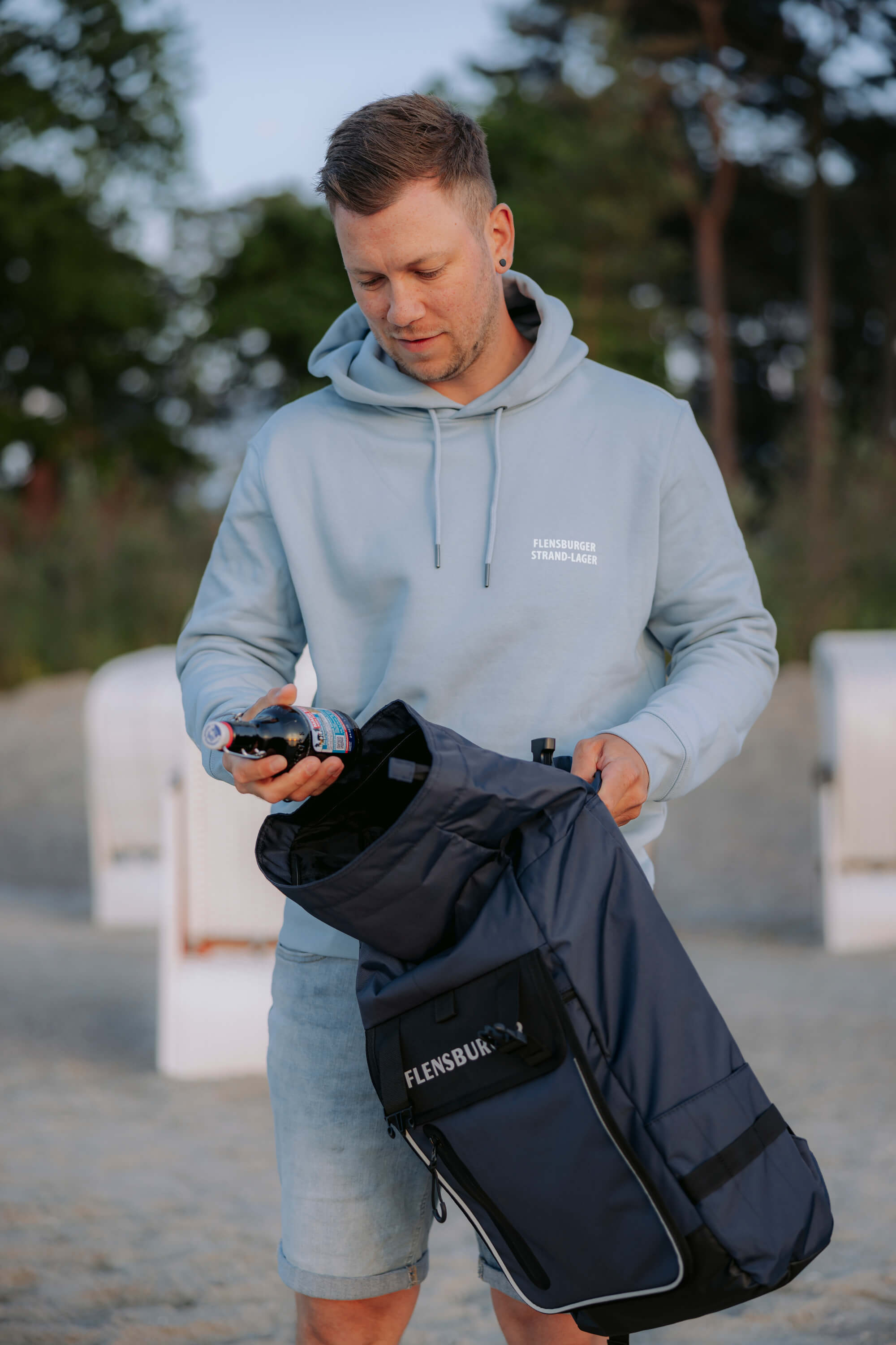 Ein Mann in hellblauem Hoodie mit der Aufschrift „Flensburger Strand-Lager“ steckt eine Flasche in den dunkelblauen Rucksack „Nordlicht Explorer“ mit der Aufschrift „Flensburg“.