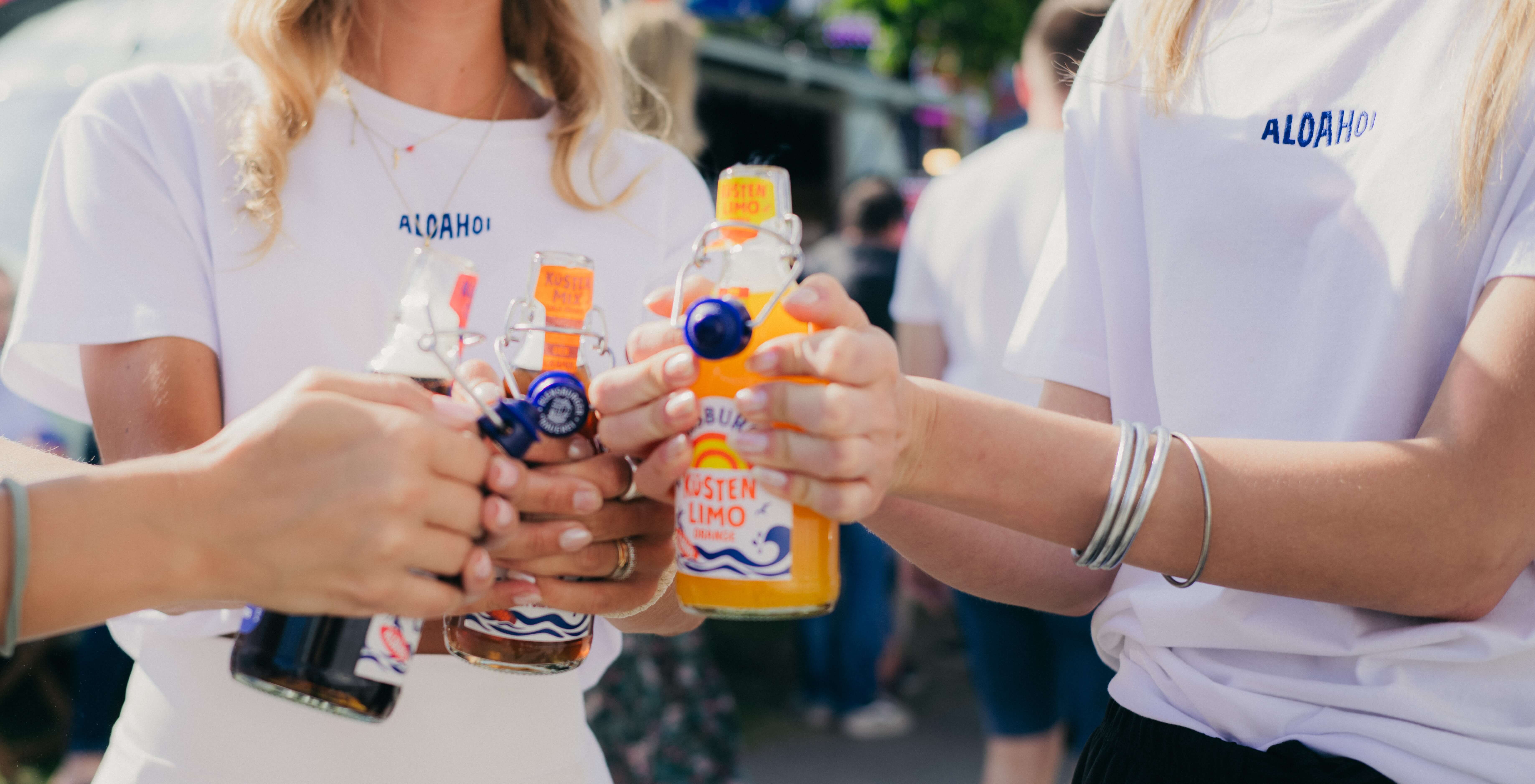 Drei Personen halten Flensburger Küstenlimos in verschiedenen Farben. Alle tragen weiße T-Shirts mit dem Aufdruck "Aloha". Ein sonniger Tag scheint herrscht.
