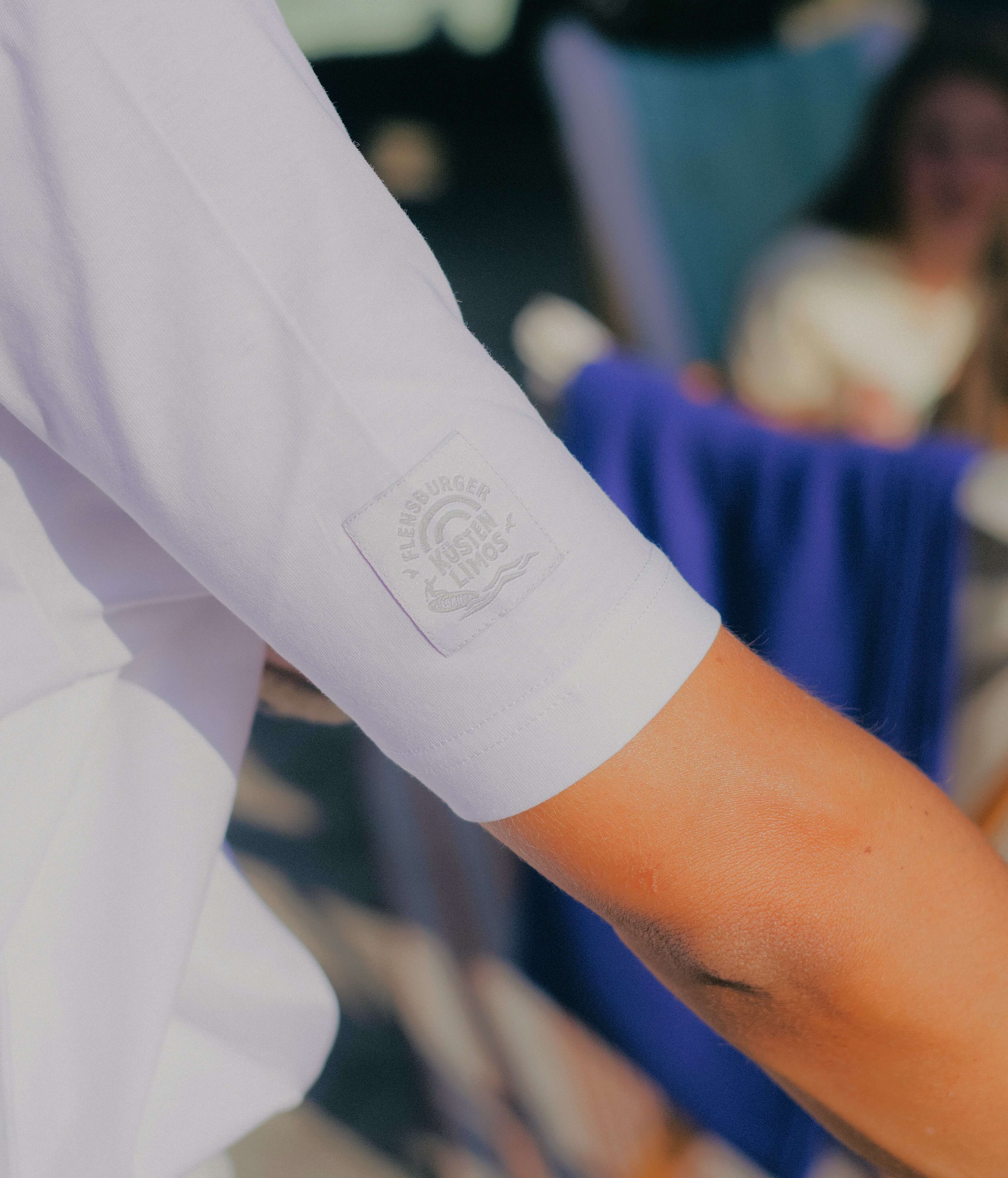 Detailansicht eines Ärmels von einem weißen Kurzarm-Shirt . Darauf ist ein geprägten Logomit der Aufschrift "Flensburger Küstenlimos" zu sehen. Im Hintergrund sind Personen und Textilien sichtbar.