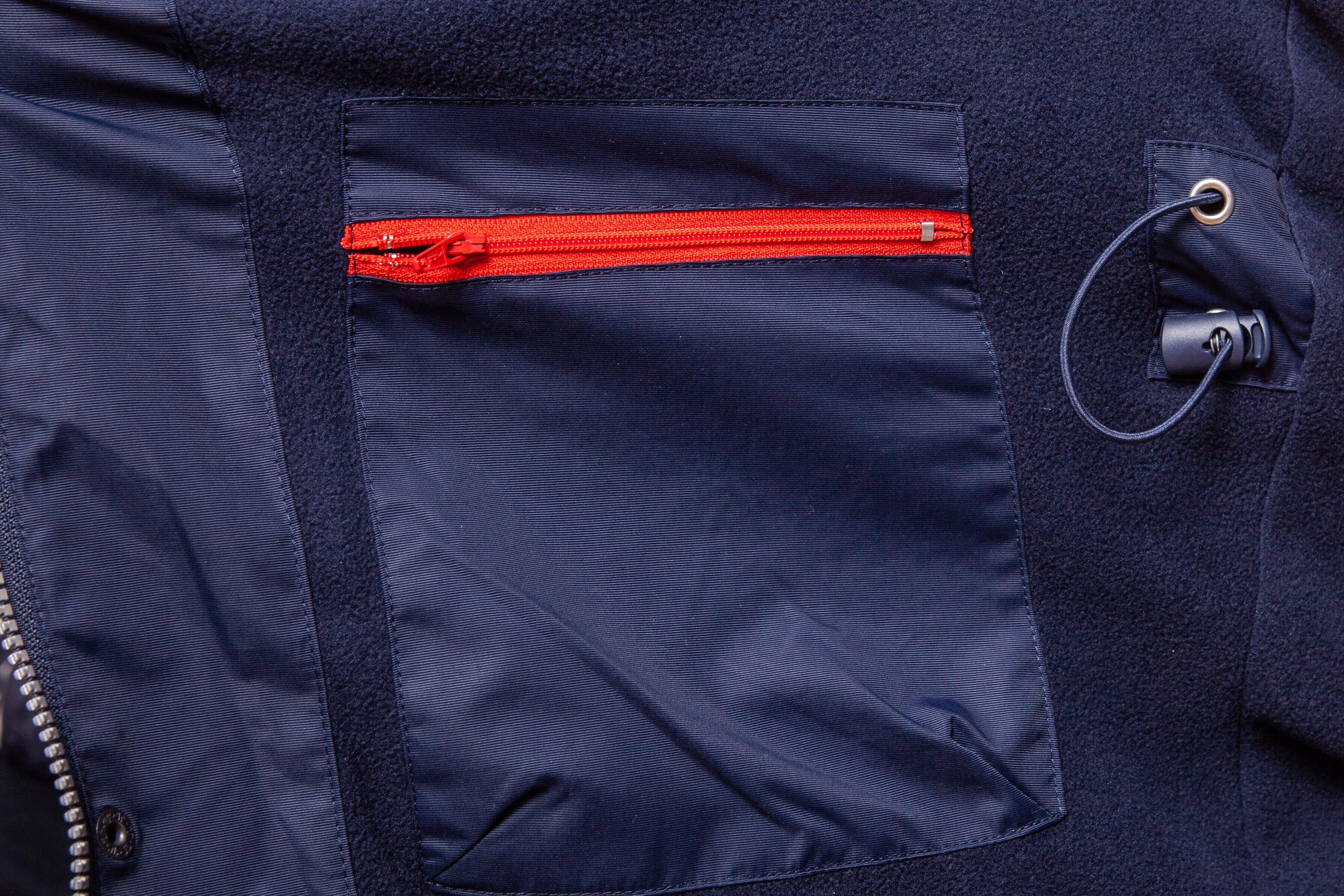 Innenseite eines navyblauen Parkas mit rotem Reißverschluss an einer Brusttasche und verstellbarem Kordelzug an der Seite. Der Stoff ist eine Kombination aus Fleece und glattem Material.