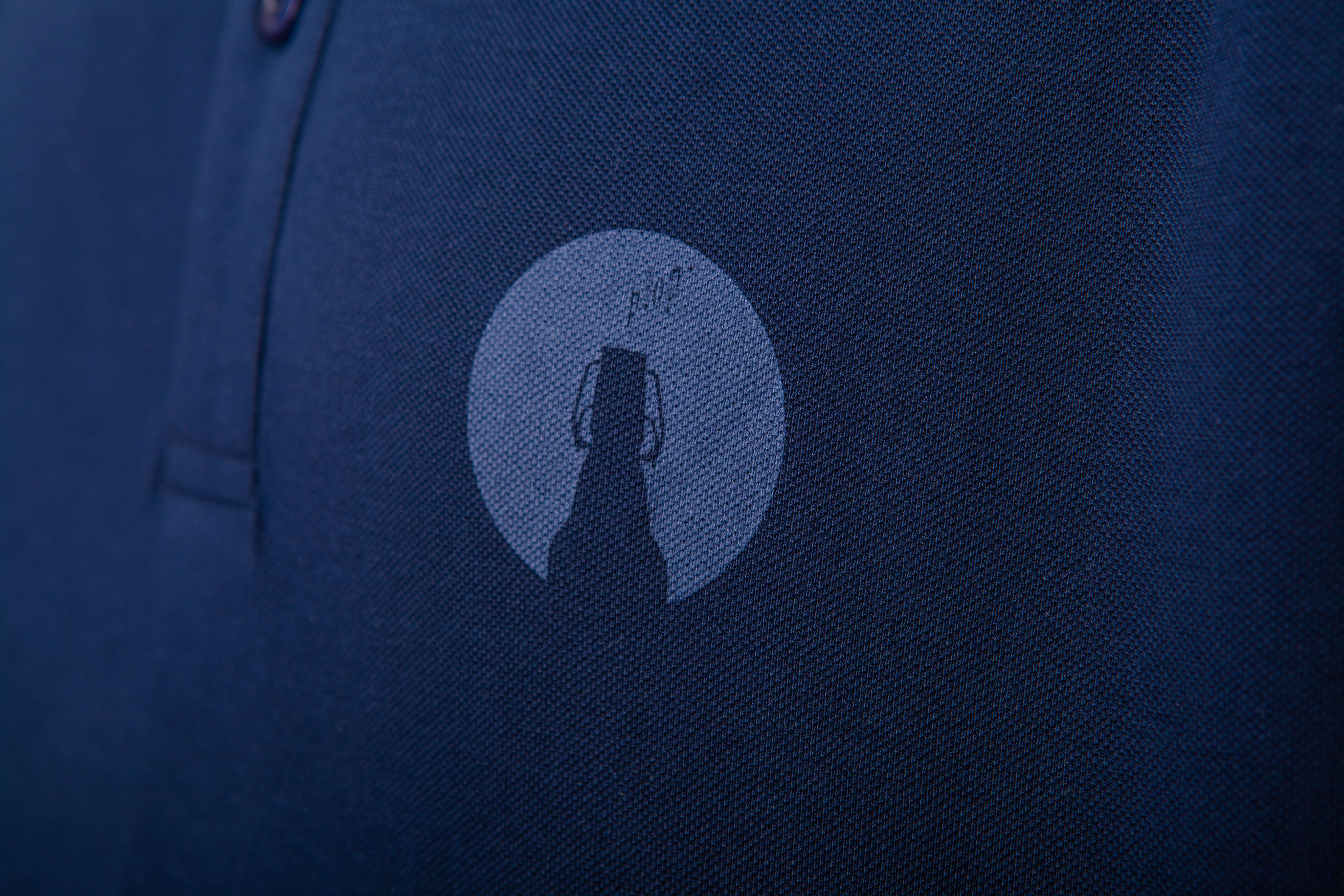 Detailansicht: Dunkelblaues Poloshirt mit einem hellblauen runden Logo auf der Brust, das eine stilisierte Bügelflasche zeigt.