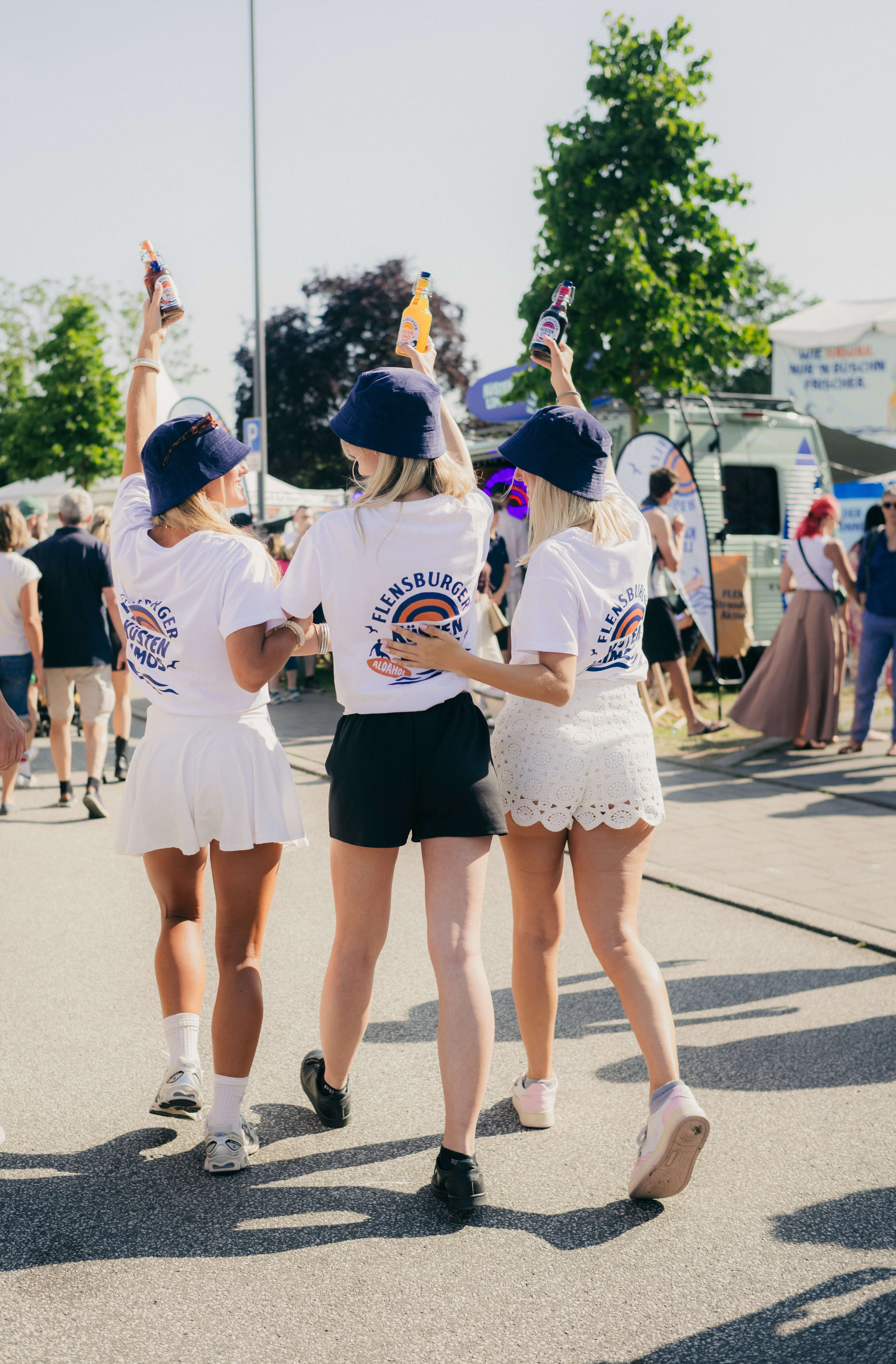 Drei Frauen tragen weiße T-Shirts mit dem Aufdruck "Flensburger Küstenlimos" und genießen den Tag in einem festlichen Ambiente. Sie heben Getränke in die Luft und lächeln.