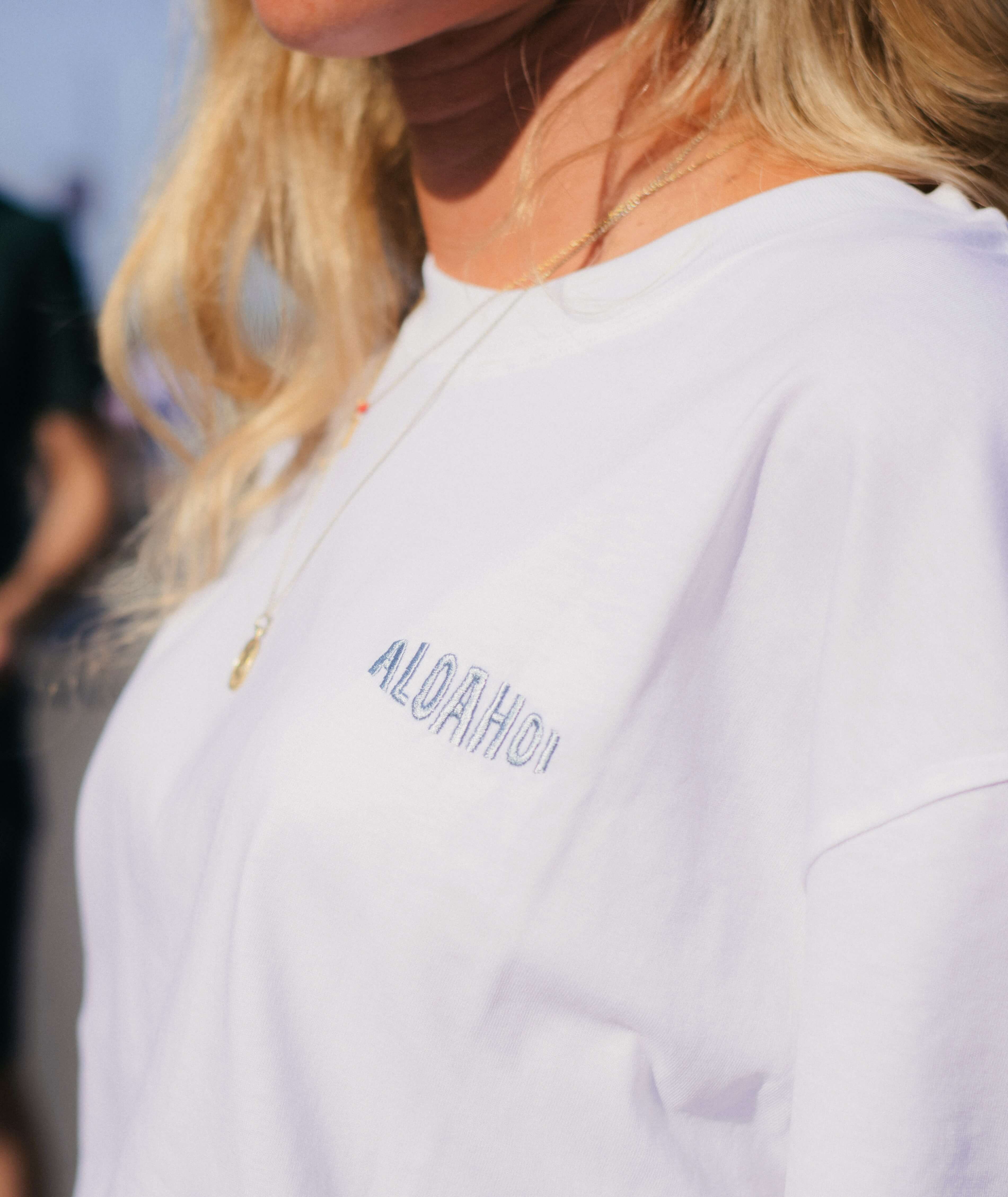 Eine Person trägt ein weißes T-Shirt mit dem aufgestickten Schriftzug "Aloahoi" in einer sanften blauen Farbe. Die Person hat blonde Haare und steht im Freien.
