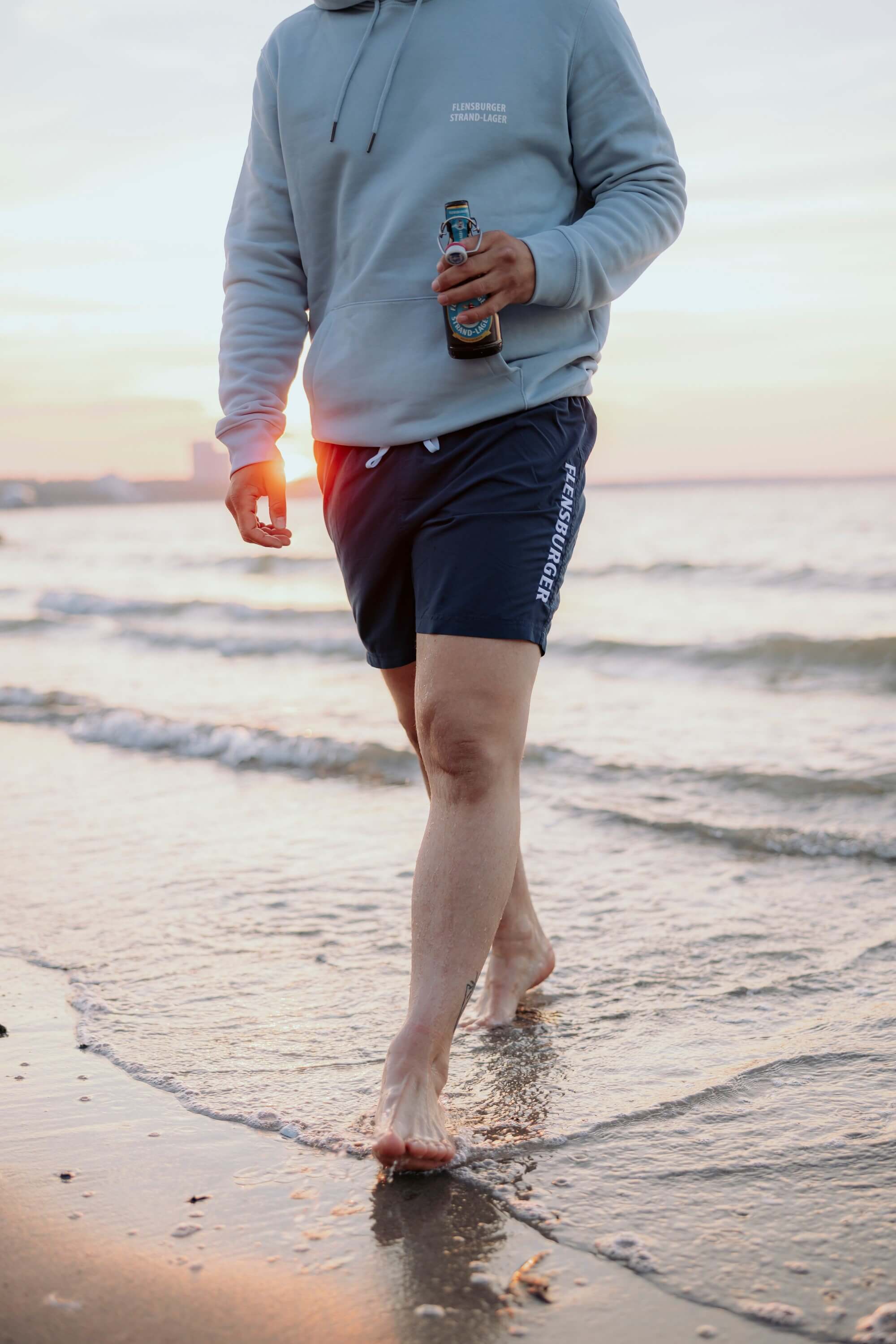 Eine Person trägt eine hellblaue Kapuzenjacke mit dem Text "FLENSBURGER STRAND-LAGER" und navyblaue Badehose mit dem Schriftzug "FLENSBURGER". Sie geht barfuß am Strand und hält eine Flasche Bier.
