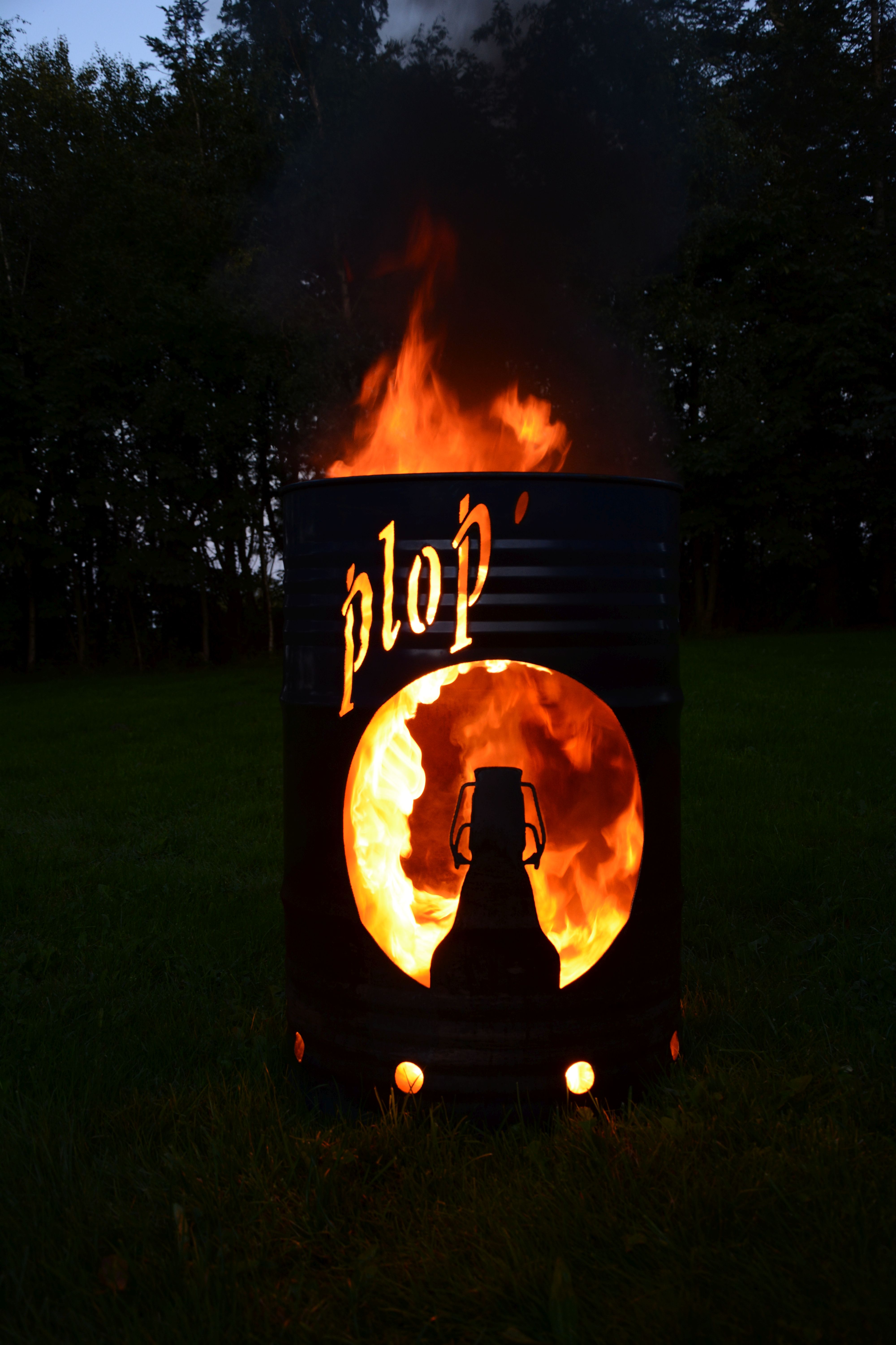 Eine Feuertonne brennt mit sichtbaren orangen Flammen. Die Tonne zeigt den Schriftzug „plop‘“ und eine ausgeschnittene Flaschenform, die von der Flamme hinterleuchtet wird.