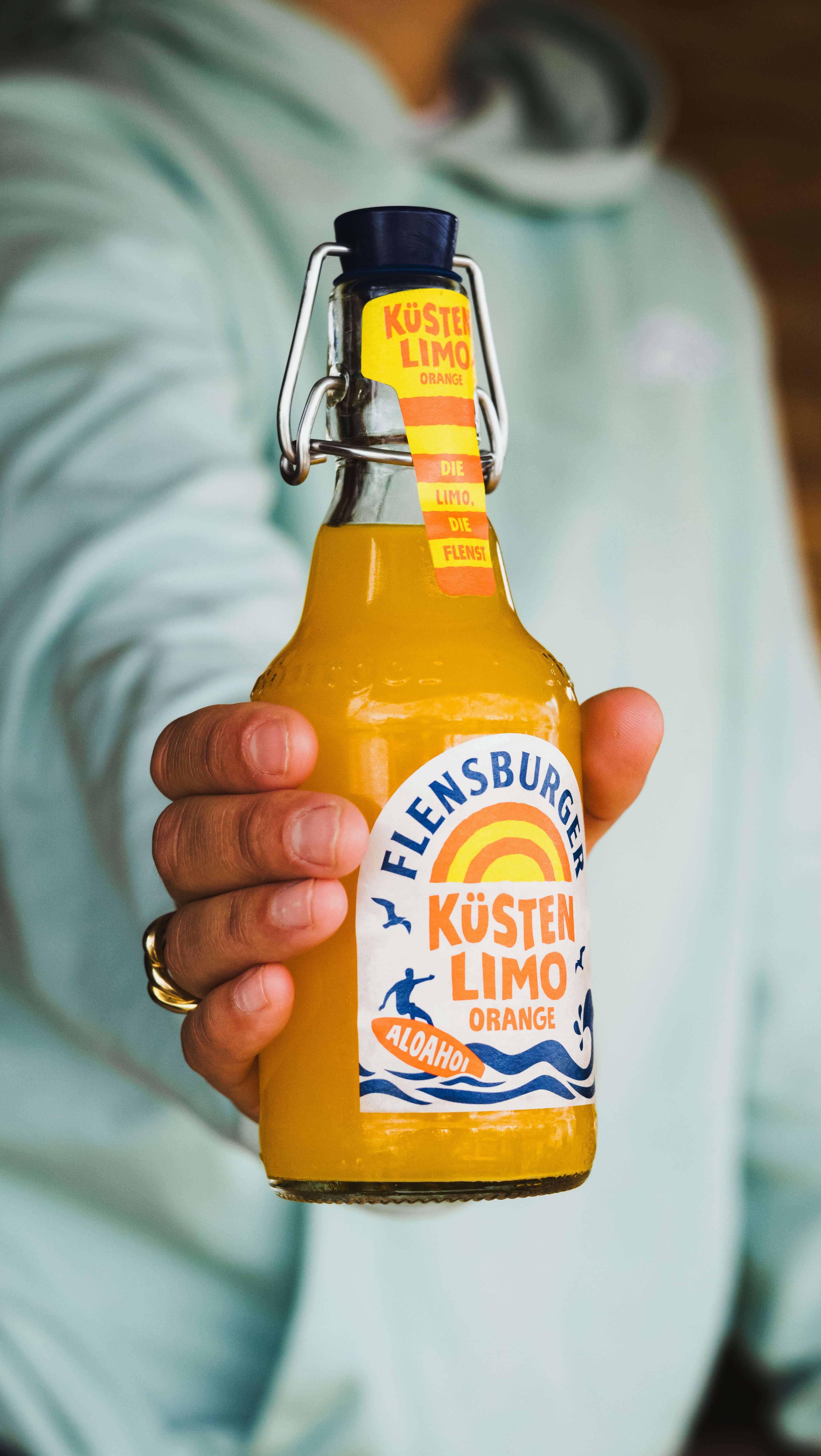 Flensburger Küstenlimo "Orange" wird von einer Person mit Hand gehalten. Die Flasche hat eine gelbe und blaue Etikettierung sowie eine fest verschlossene Kappe.