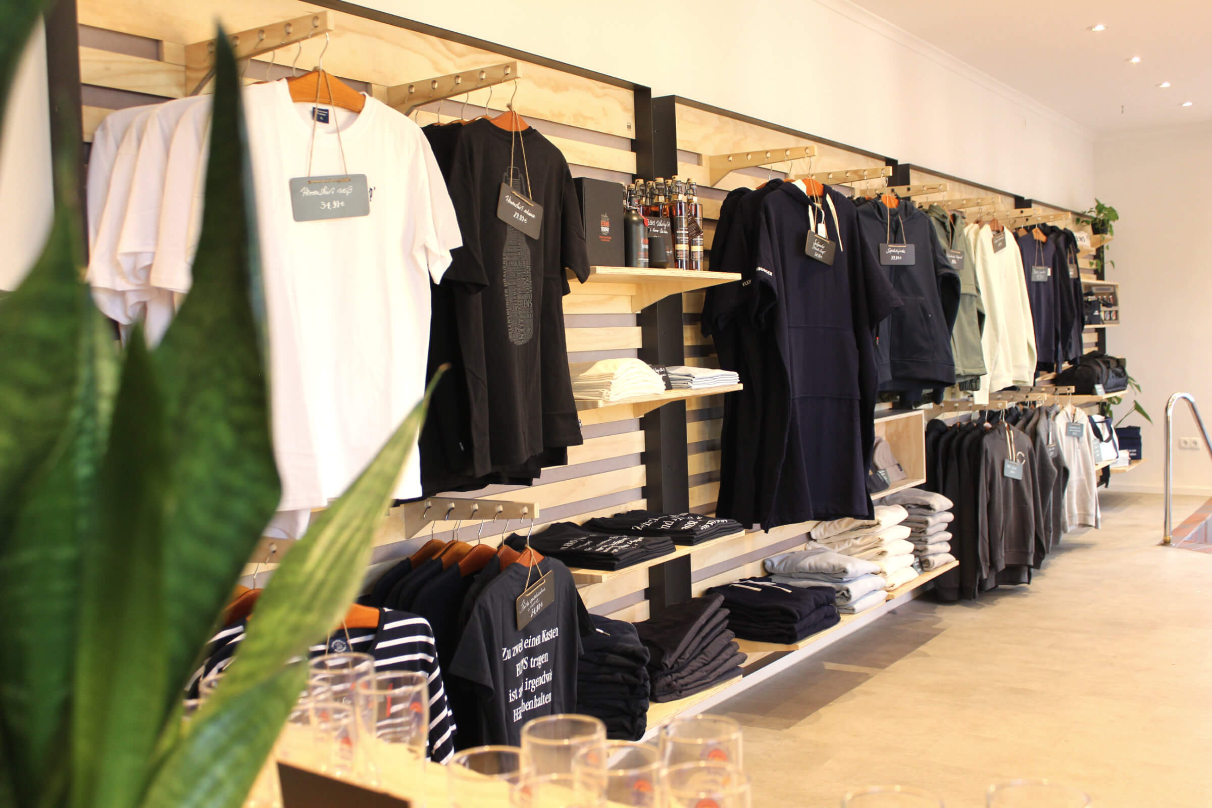 An der Wand hängen verschiedene Kleidungsstücke, vor allem T-Shirts in verschiedenen Farben (weiß, schwarz, dunkelblau). Darunter sind weitere gefaltete Kleidungsstücke ordentlich auf Regalen gestapelt.