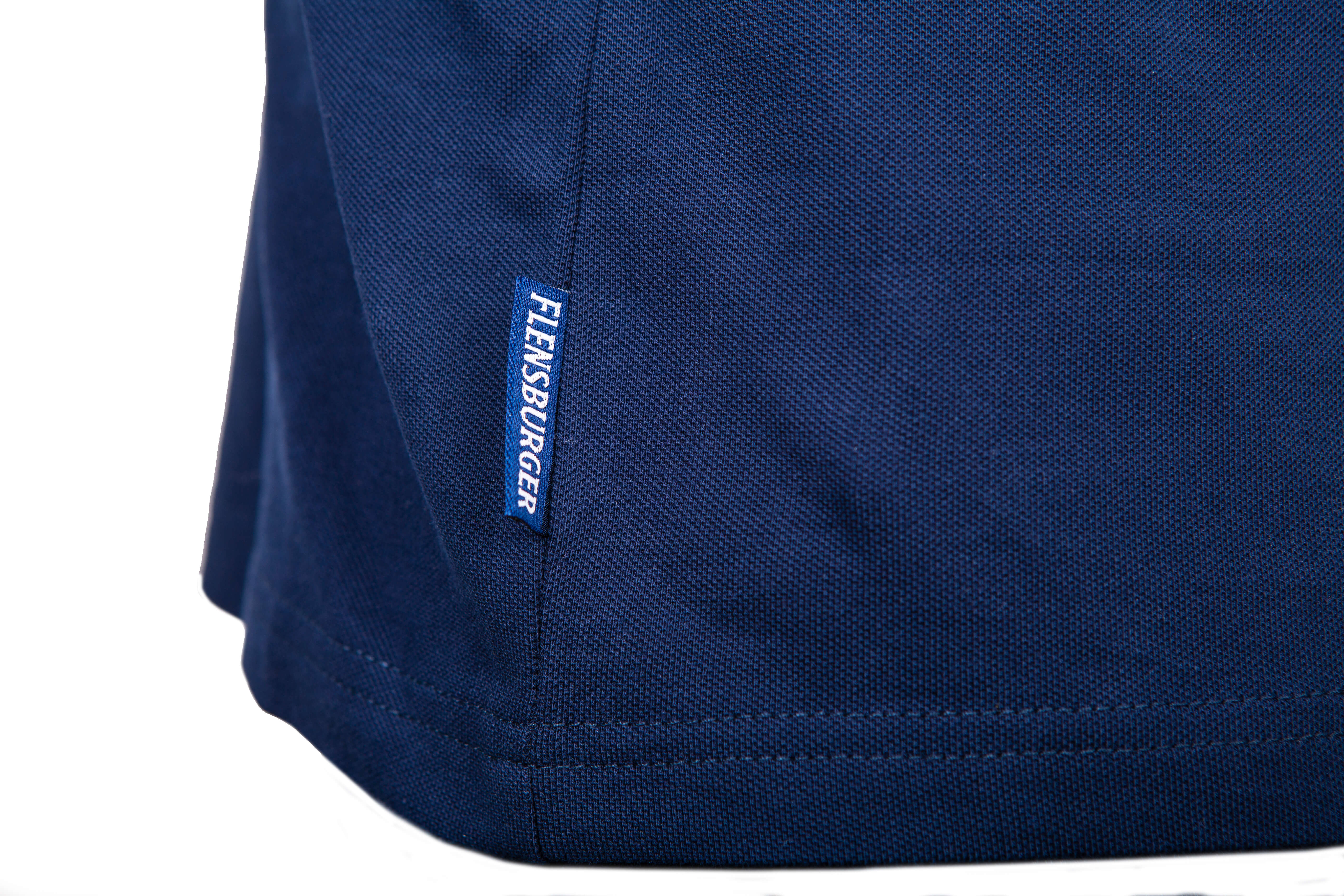 Detailansicht: Dunkelblaues Poloshirt aus Baumwolle mit einem kleinen weißen Schriftzug “FLENSBURGER” auf blauem Etikett an der Seitennaht.