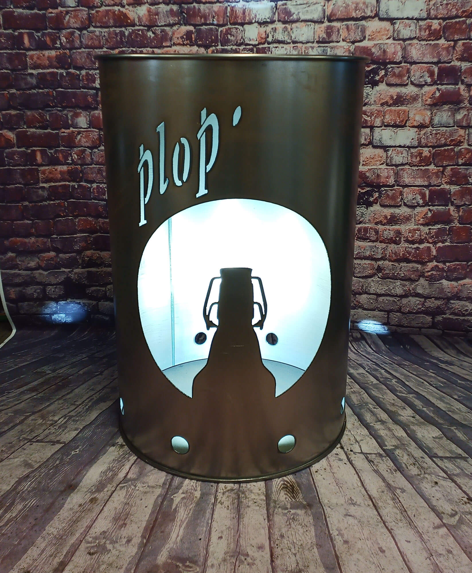 Metallzylinder mit eingeprägtem Schriftzug „plop’“ zeigt eine ausgeschnittene Flaschenkontur, die von innen beleuchtet wird.