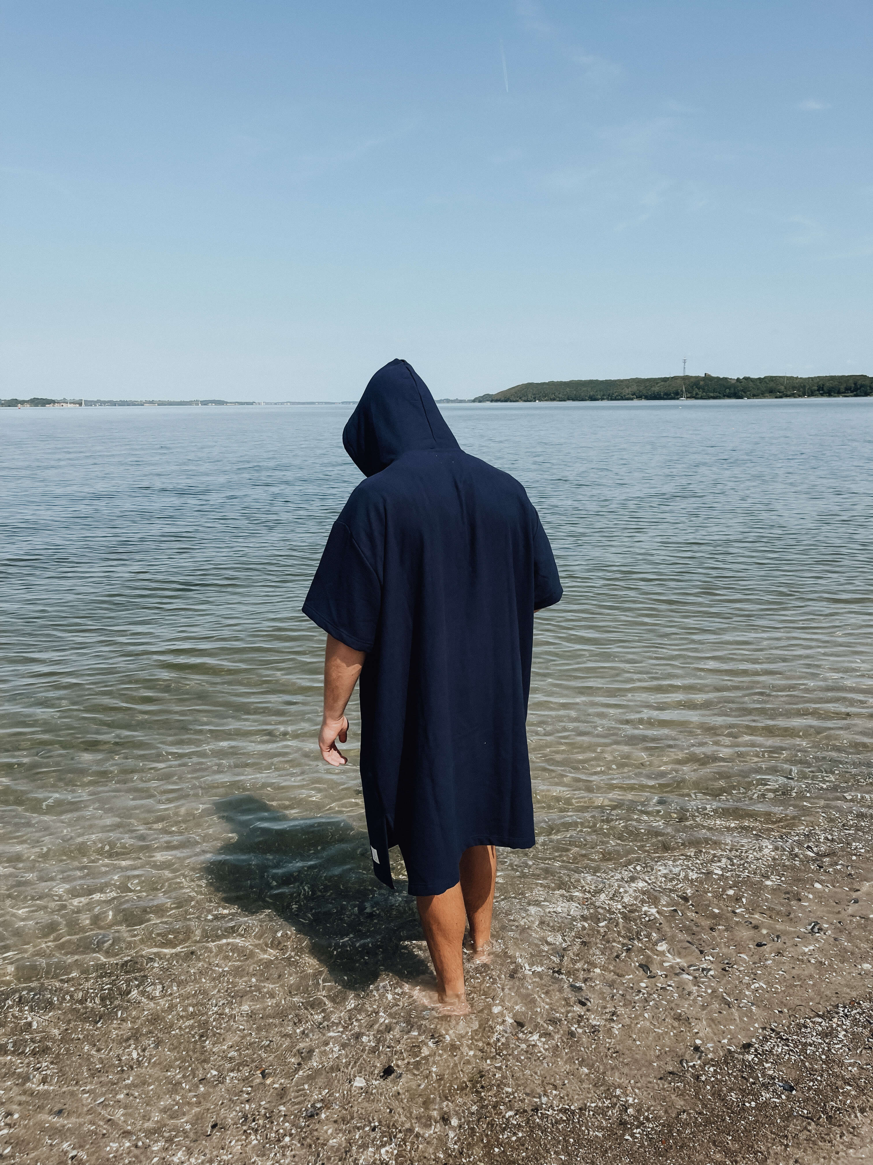 Eine Person trägt den blauen Strand-Poncho "Strand-Lager" und steht mit nackten Füßen im flachen Wasser an einem Strand. Der Poncho hat kurze Ärmel und eine Kapuze.