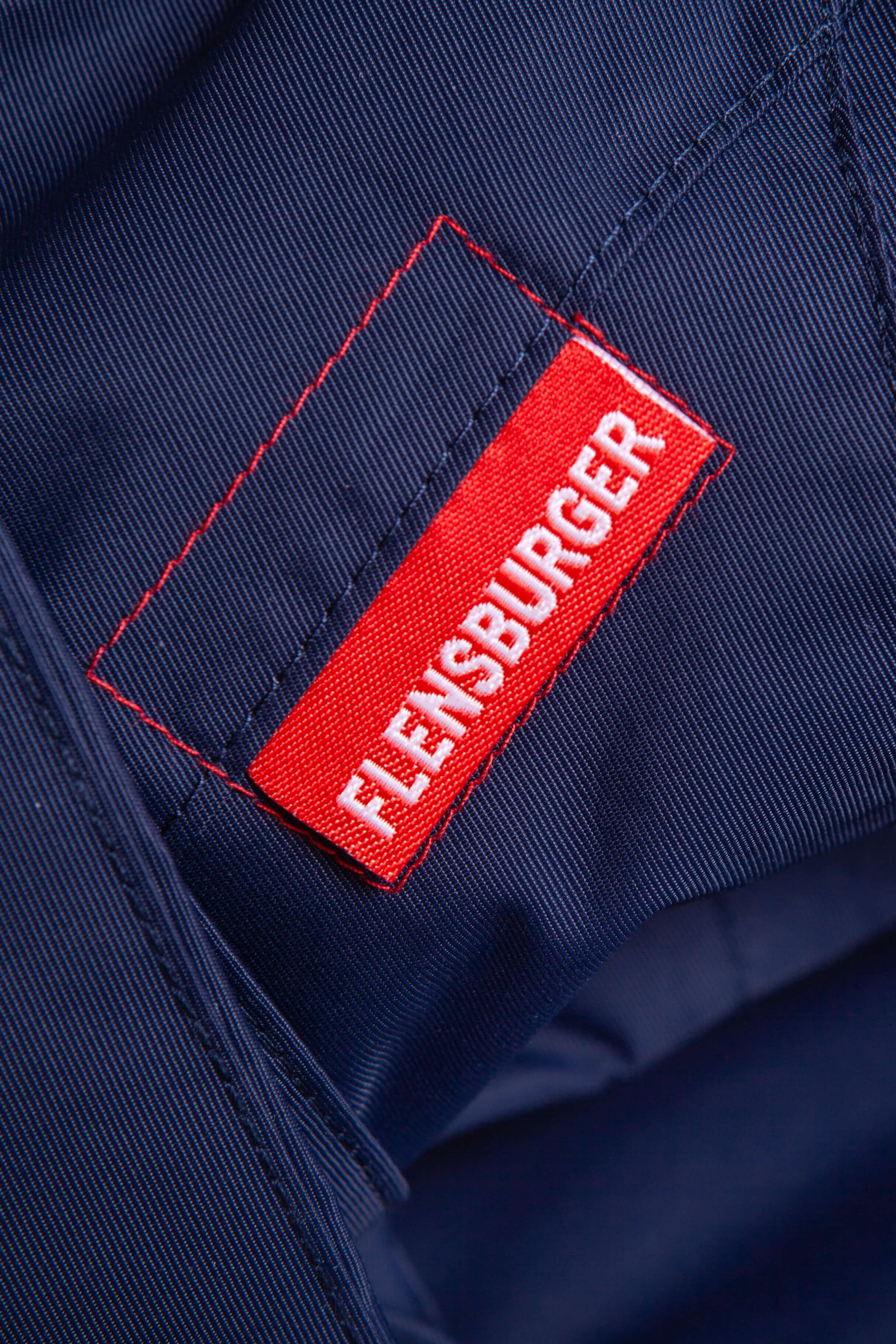 Rotes Etikett mit weißer Aufschrift "FLENSBURGER" auf navyblauem Parka-Stoff mit sichtbaren roten und schwarzen Nähten.