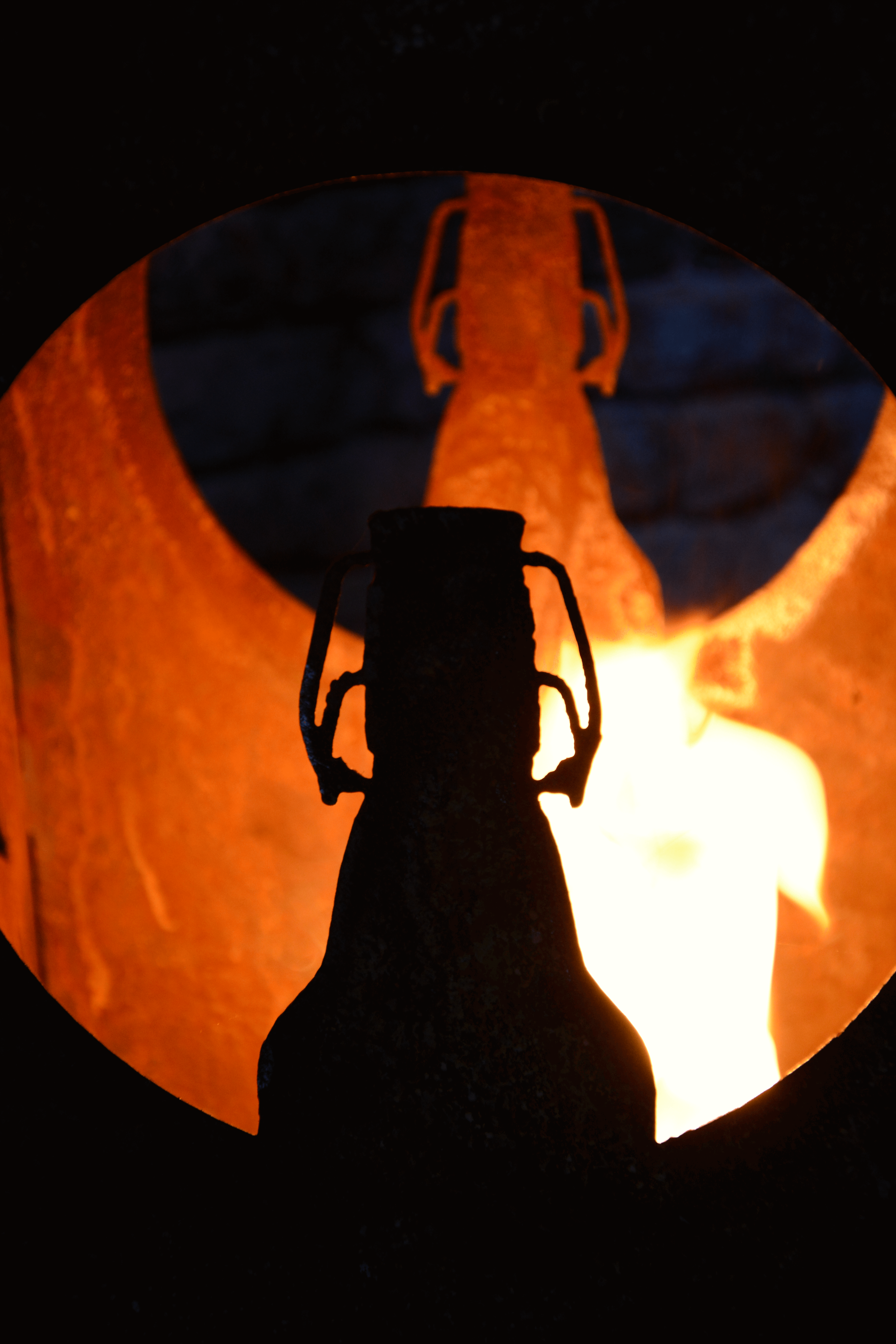 Ein silberner Feuerkorb mit einer Flasche in der Mitte, die vor einer brennenden Flamme als dunkle Silhouette zu sehen ist. Das Metall ist rund und offen gestaltet.