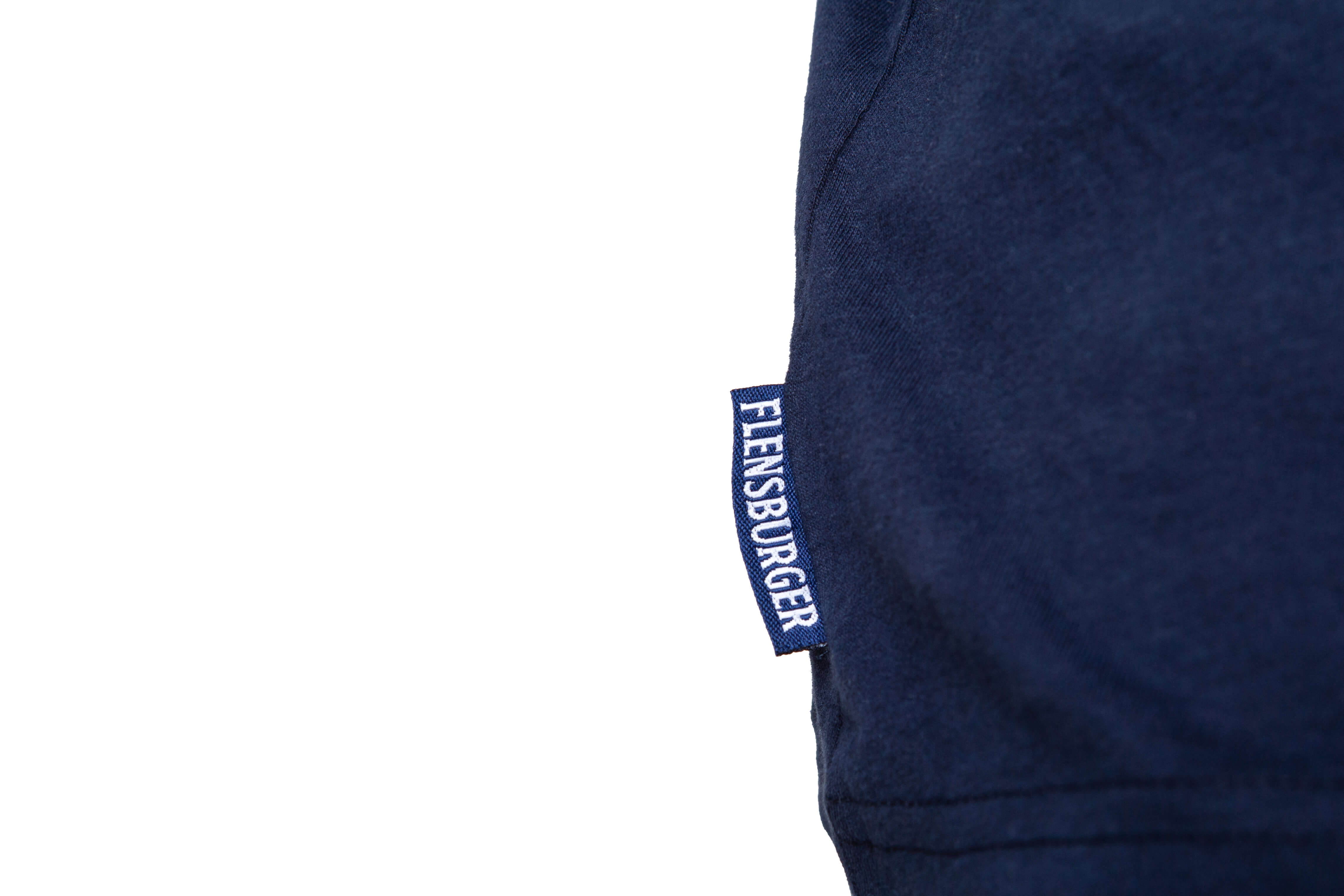 Detailansicht: Blaues T-Shirt mit einem Etikett an der Seitennaht, auf dem in weißen Buchstaben „FLENSBURGER“ steht. Das Material wirkt weich und ist aus Baumwolle.