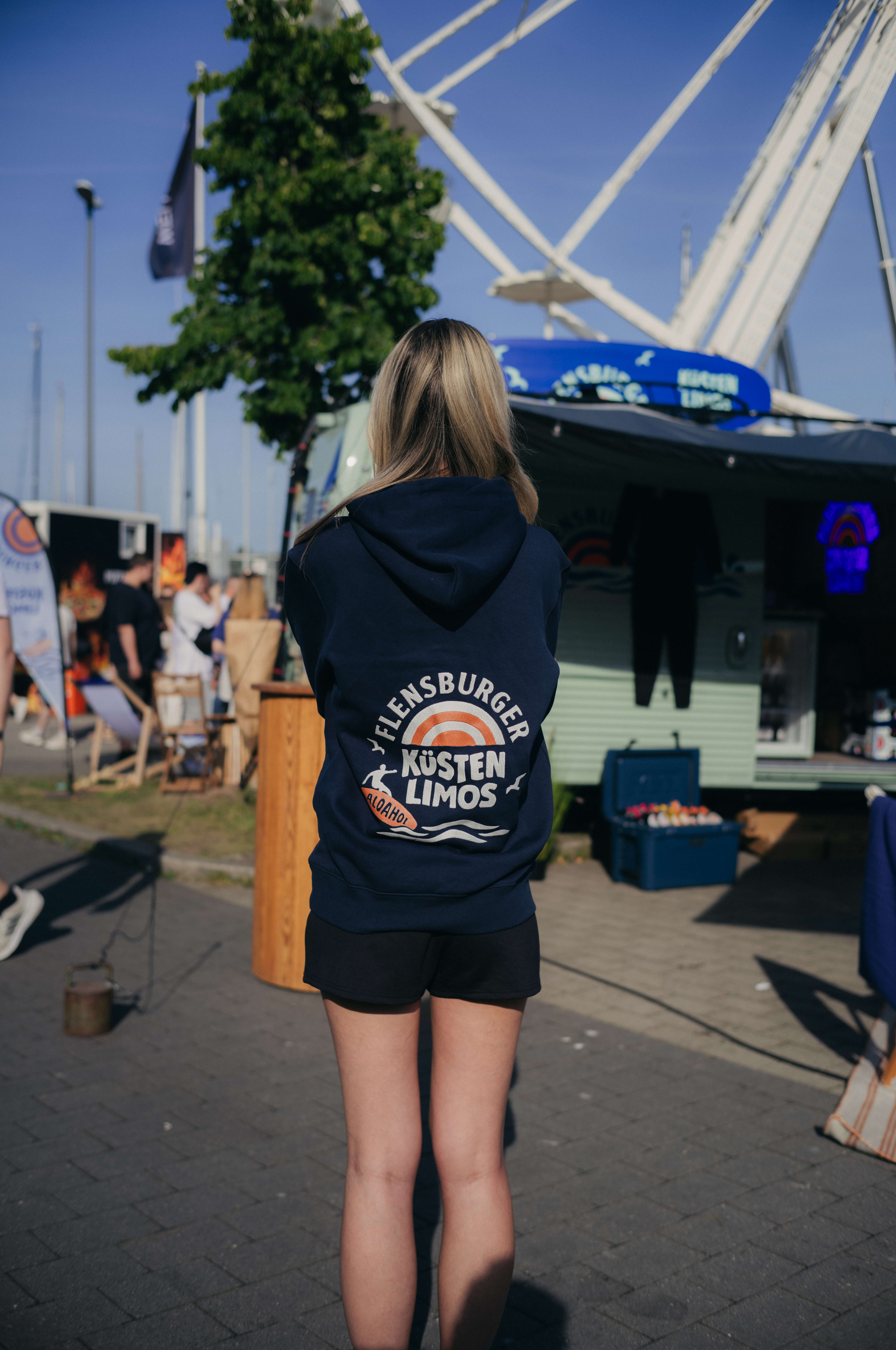 Eine Person trägt einen navy blauen Hoodie mit dem Aufdruck "Flensburger Küstenlimos". Sie steht vor einem Verkaufsstand mit lebhaften Farben und einem großen Riesenrad im Hintergrund.