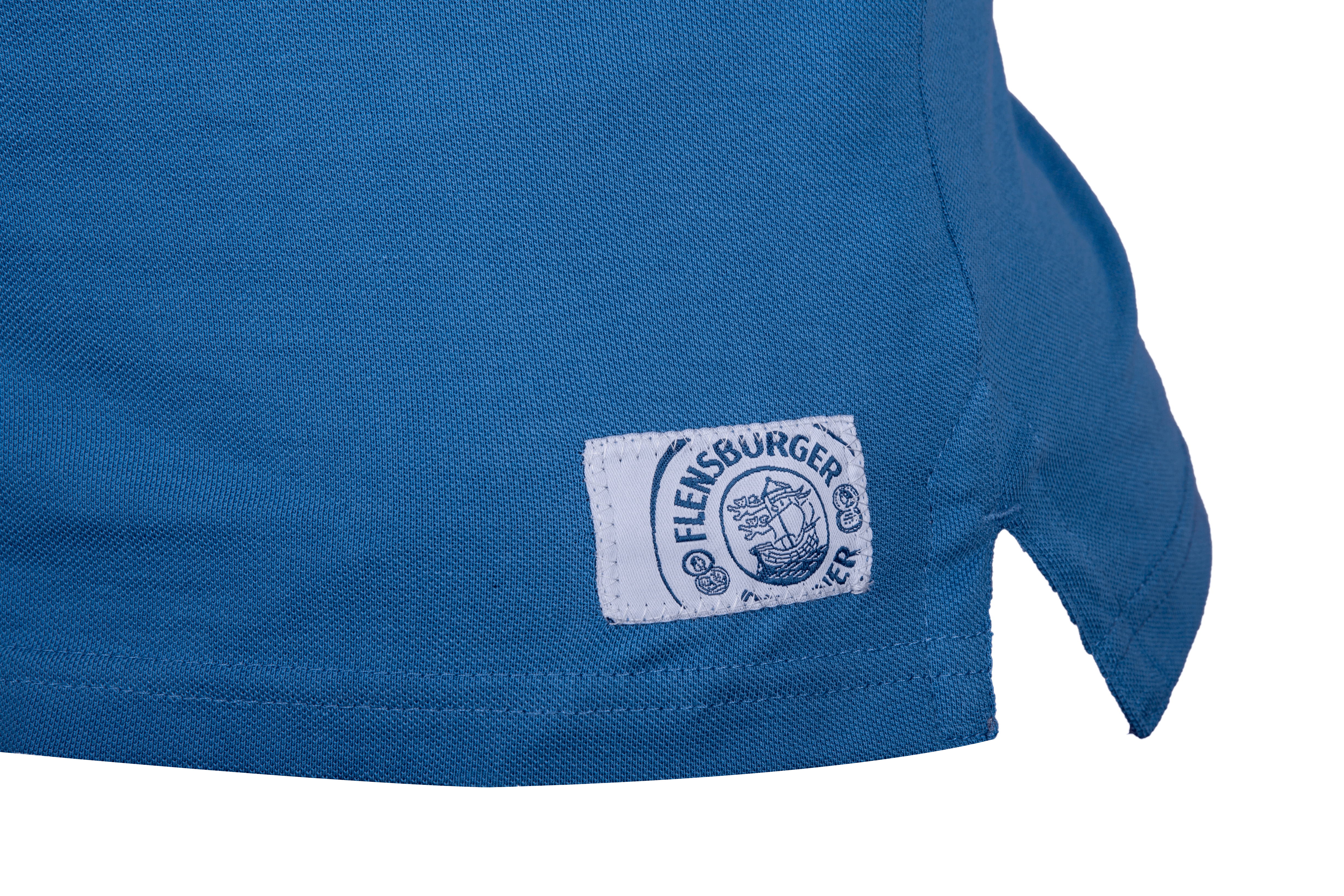 Detailansicht: Blaues Damen Poloshirt mit weißem Stofflabel am Saum. Auf dem Label steht „Flensburger“ mit einem Segelschiff-Logo in dunkelblau.
