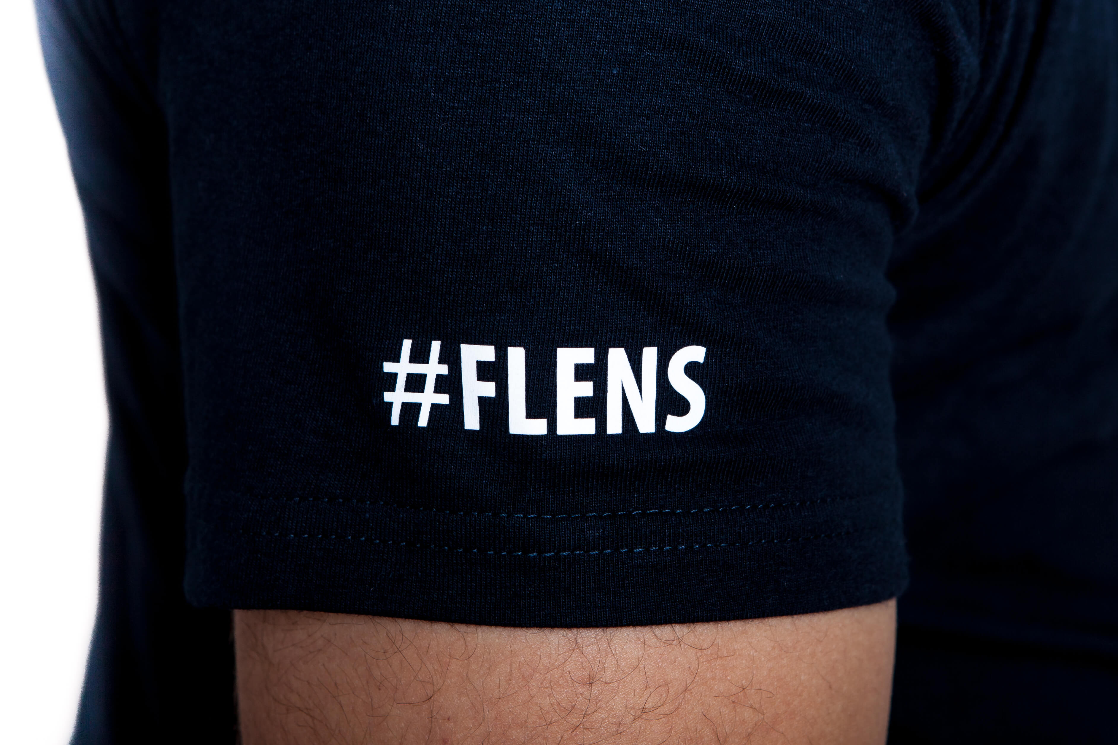 Detailaufnahme: Schwarzes T-Shirt mit dem weißen Schriftzug „#FLENS“ auf dem linken kurzen Ärmel aus 100% Baumwolle.