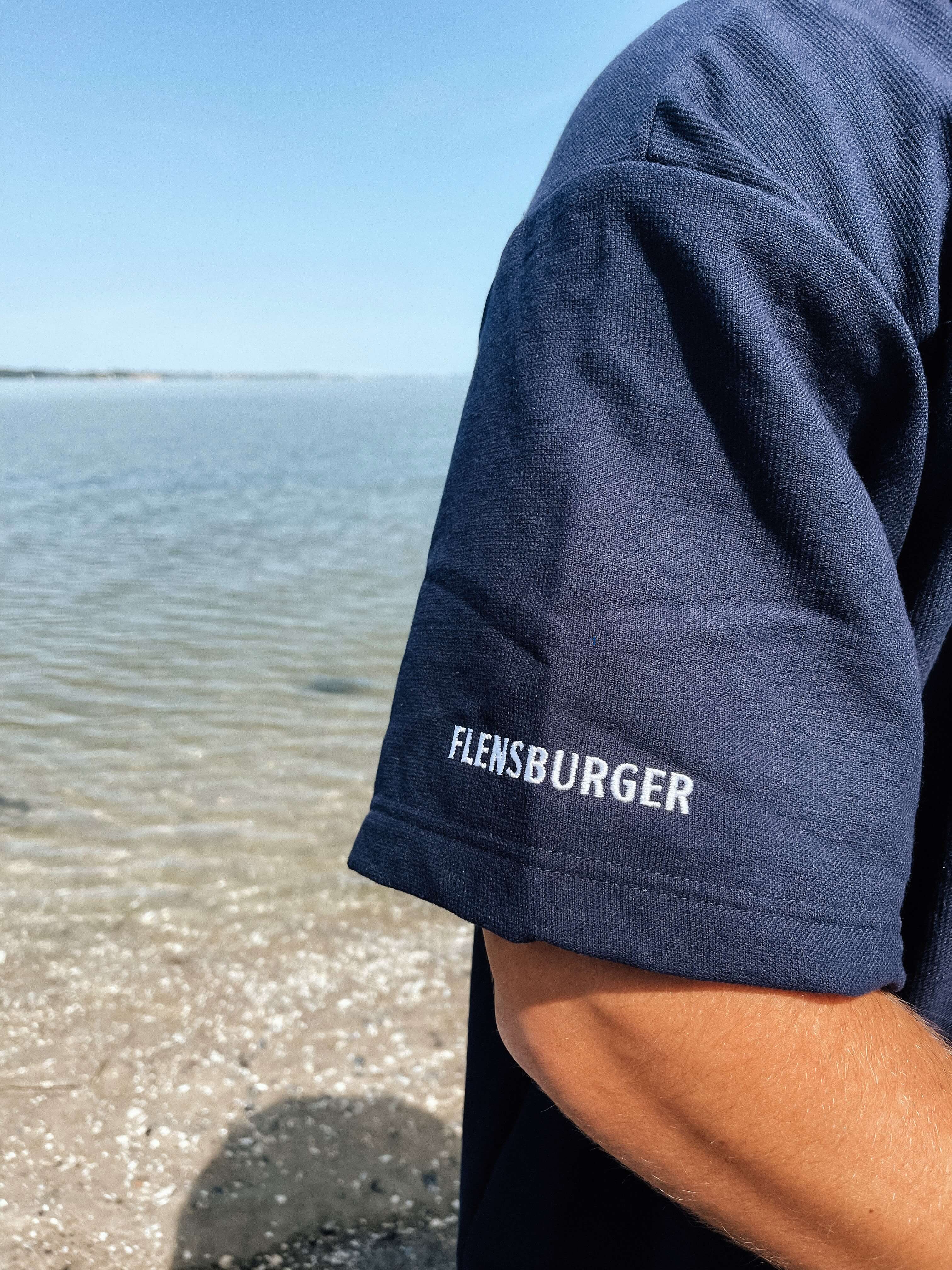 Kurzarm eines blauen Baumwoll-T-Shirts mit dem weißen Schriftzug "FLENSBURGER" am Ärmel vor einem Strand mit Wasser und klarem Himmel.