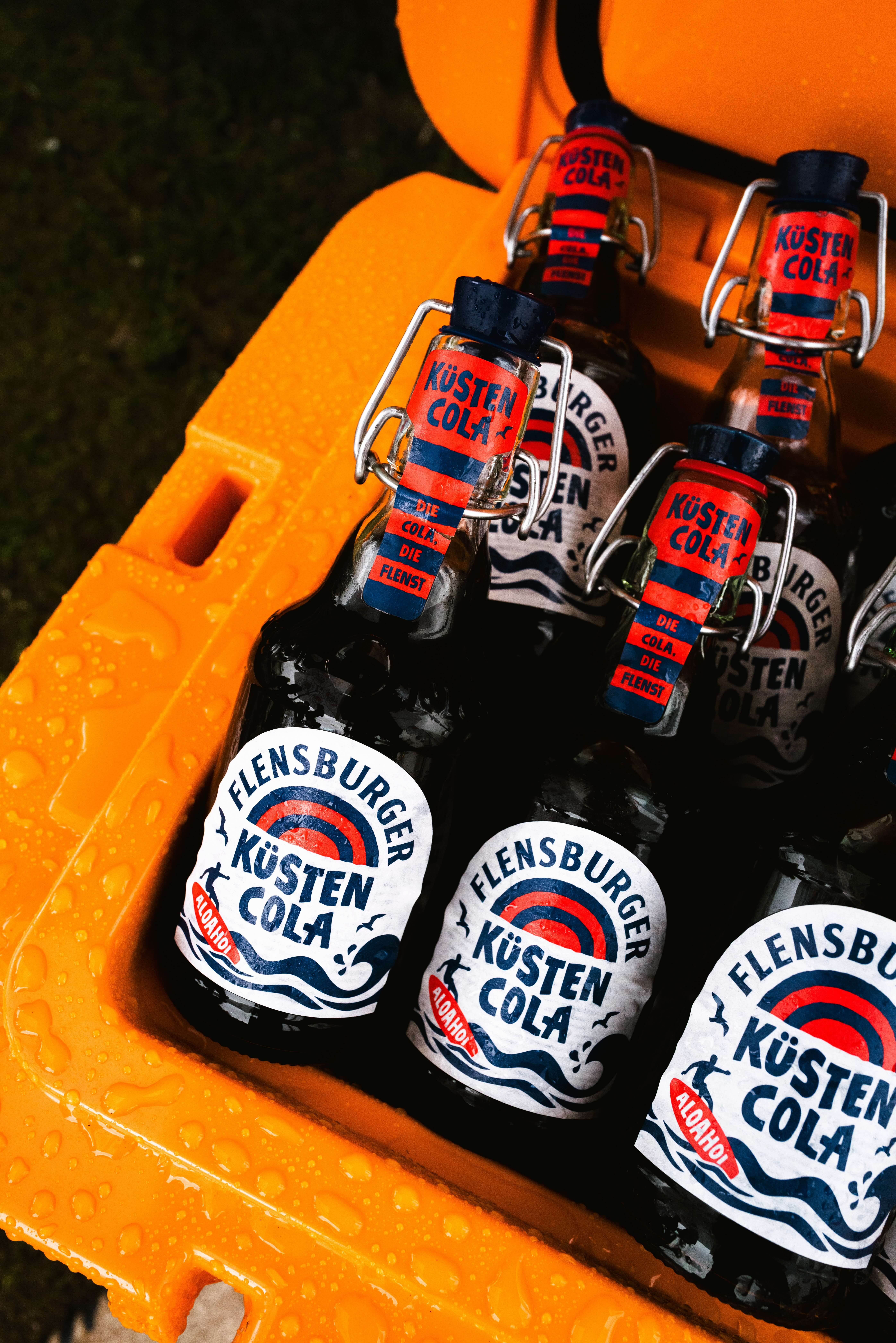 Flensburger Küstencola  Sechs Flensburger Küstencola-Flaschen in einer orangefarbenen Kiste. Die Flaschen besitzen einen Bügelverschluss und sind mit einem Etikett versehen, das den Namen und die Beschreibung der Cola anzeigt.