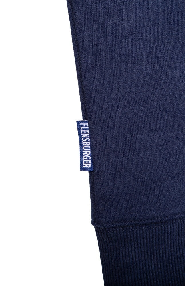 Ärmelsaum eines blauen Langarm-Pullovers mit geripptem Bündchen. Am Ärmel ist ein kleines dunkelblaues Stoffetikett mit der weißen Aufschrift „FLENSBURGER“ angenäht.