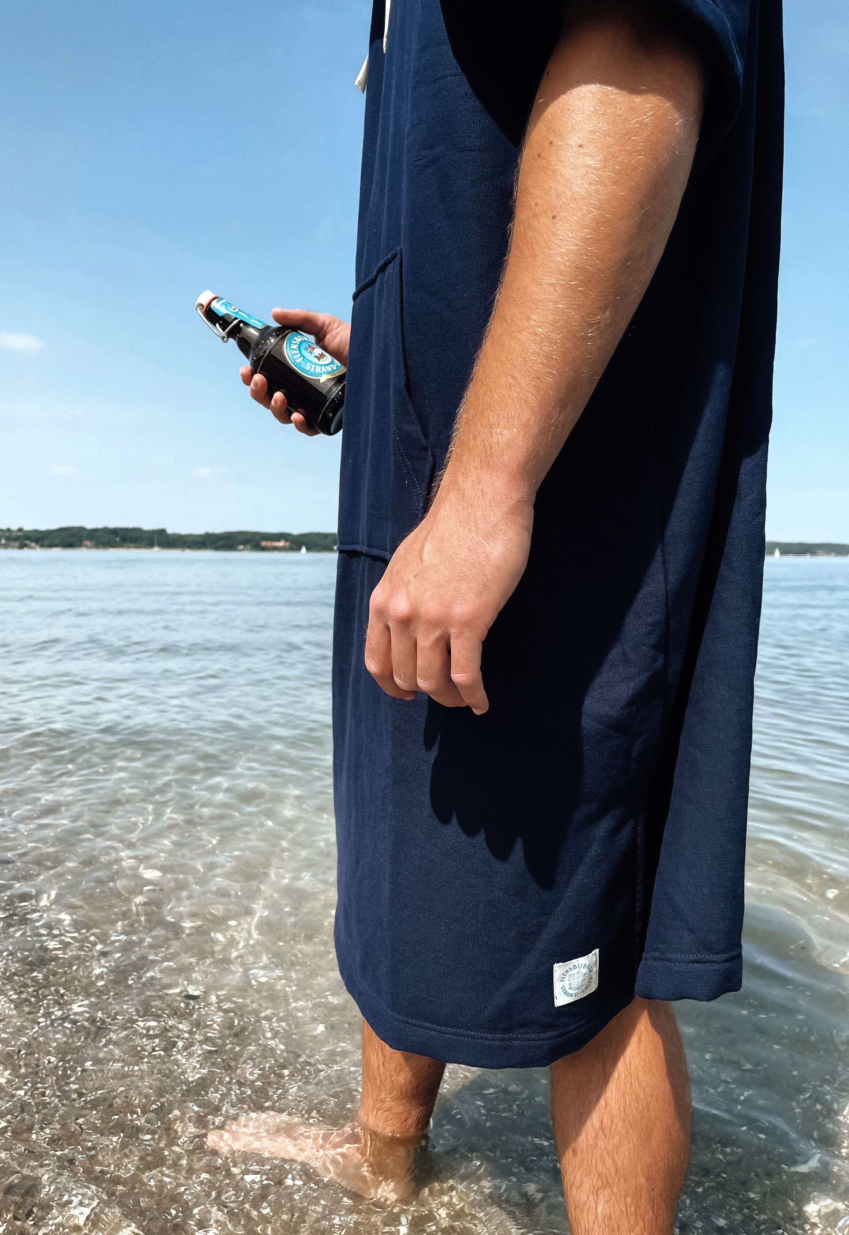 Person steht mit nackten Füßen im flachen Wasser und trägt einen blauen Strand-Poncho mit kurzem Arm. Die Hand hält eine dunkle Flasche mit blau-weißem Etikett.