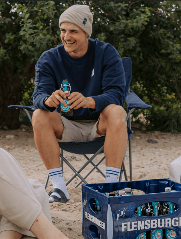 Ein Mann trägt einen weißen Strickmütze und einen navyblauen Pullover. Er sitzt auf einem Campingstuhl, hält eine Bierflasche der Marke Flensburger und lacht. Vor ihm steht eine blaue Flensburger-Kiste mit weiteren Flaschen.